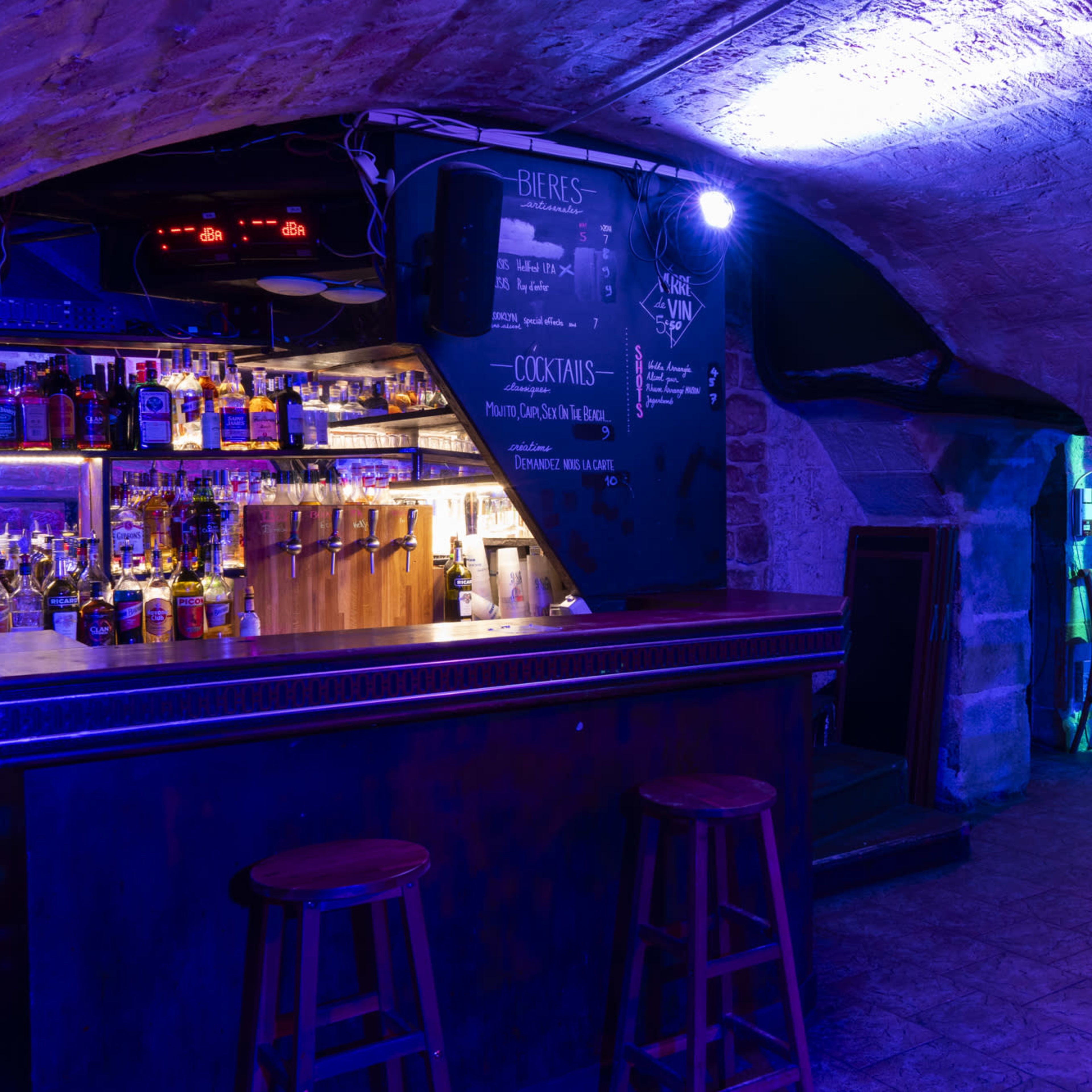 A dimly lit bar area with a wooden counter, shelves of liquor in the background, and blue lighting illuminating the space.