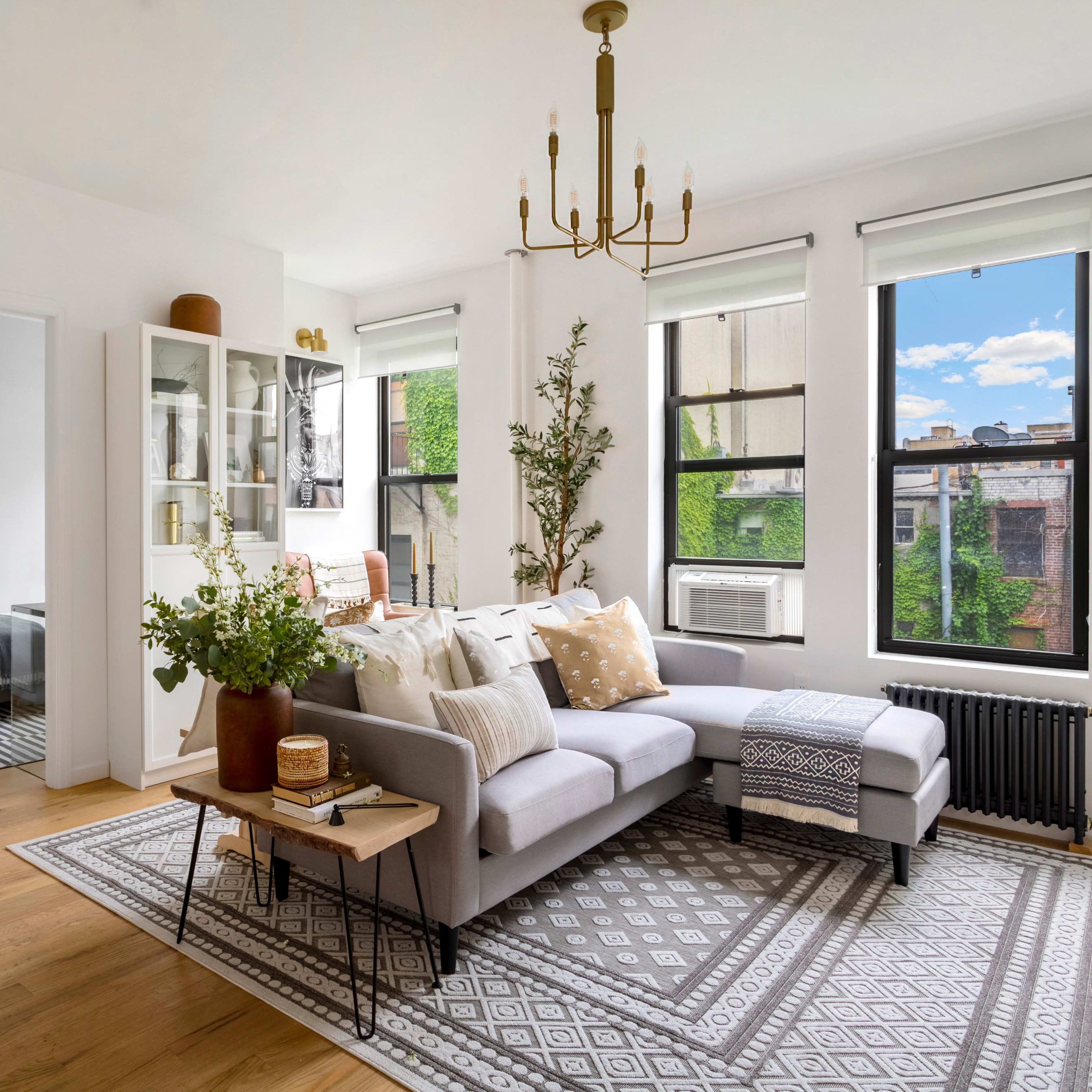 The living room features a gray sectional sofa, a decorative rug, large windows with greenery outside, and a doorway leading to another room.