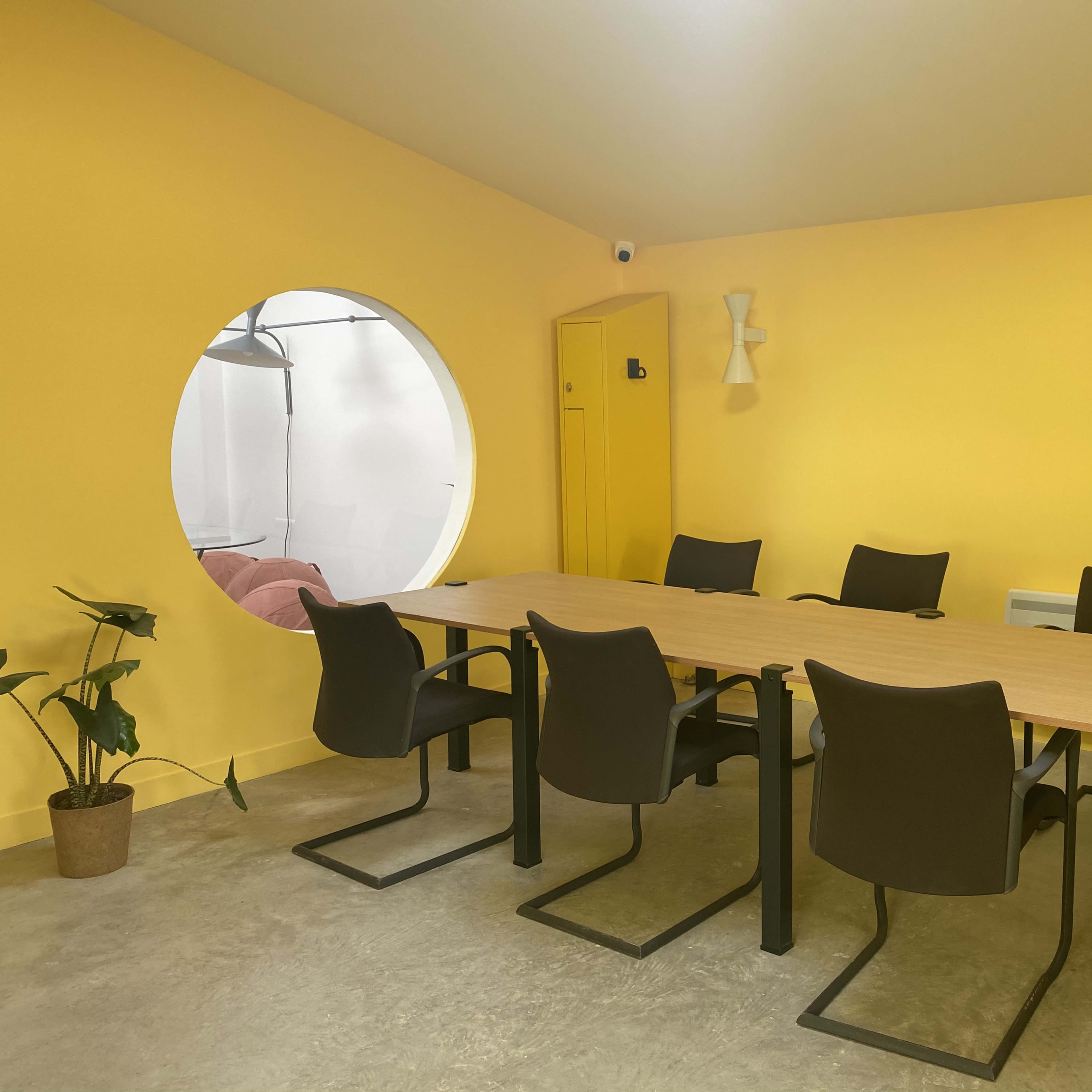 A meeting room with a large yellow wall, a wooden table surrounded by six black chairs, and a circular window leading to another room.