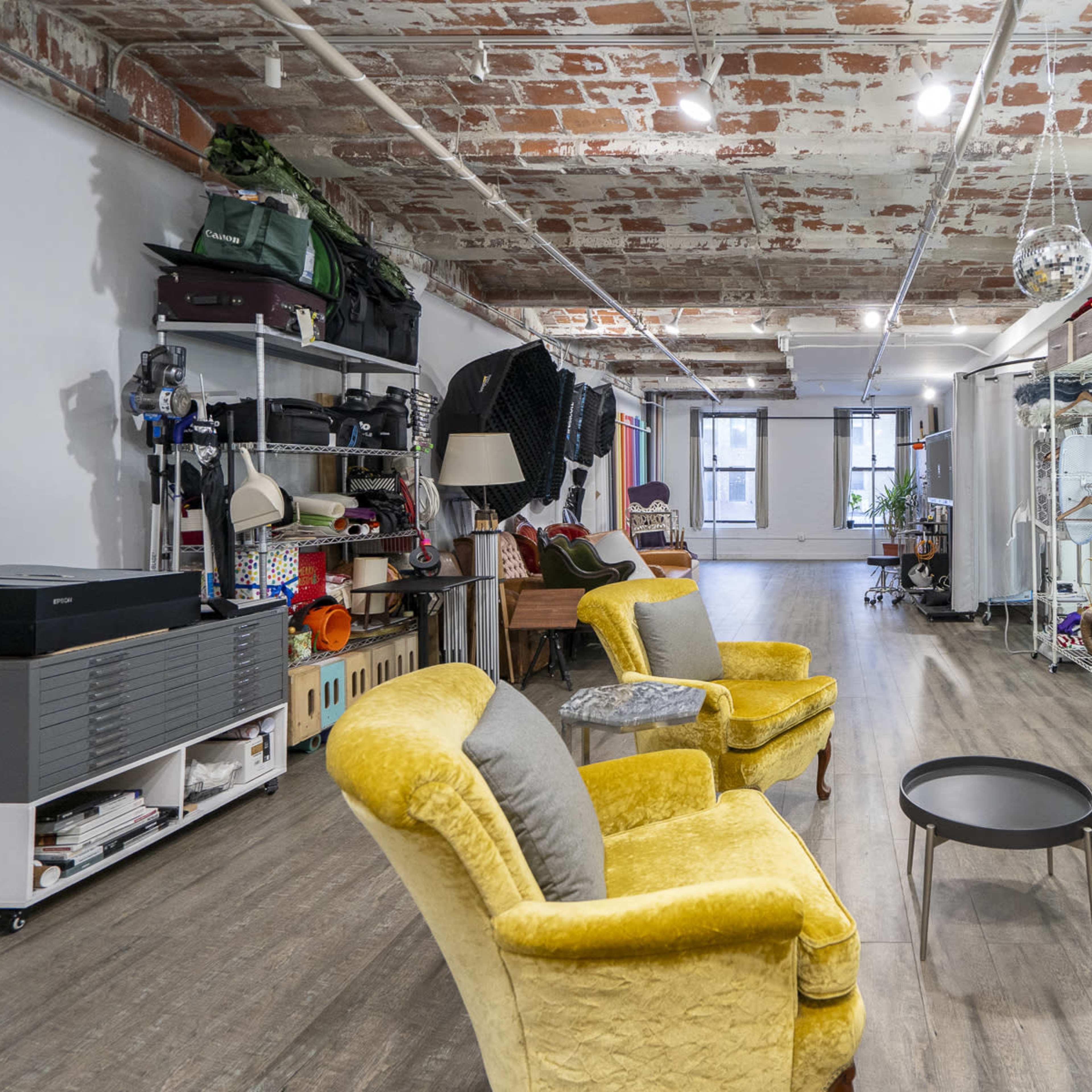 An open, well-lit interior space featuring a mix of vintage and modern furnishings, with a yellow armchair, a brown leather sofa, and various storage items along the walls.
