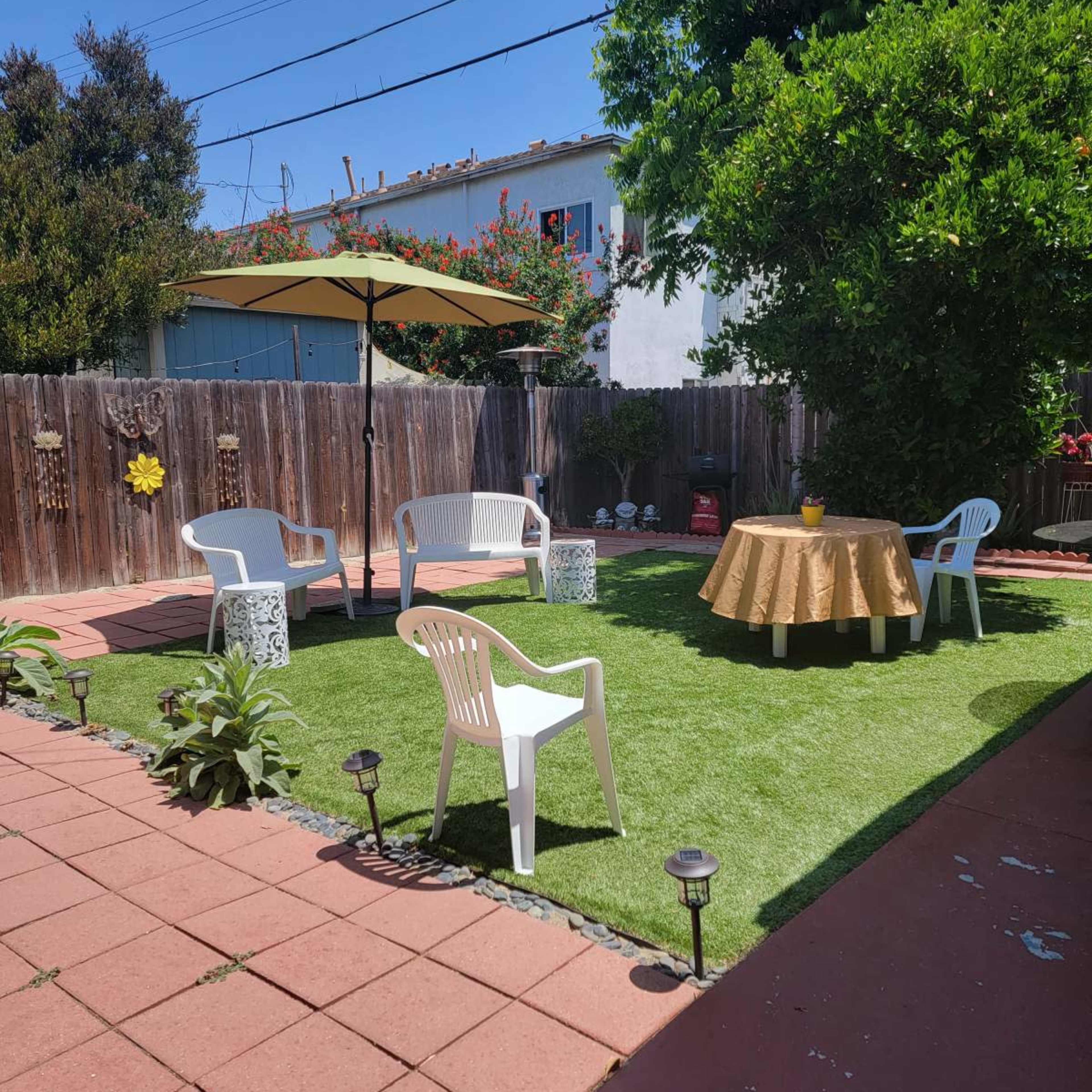 A backyard with green artificial grass, a round table covered with a gold tablecloth, and white chairs arranged under a large umbrella.