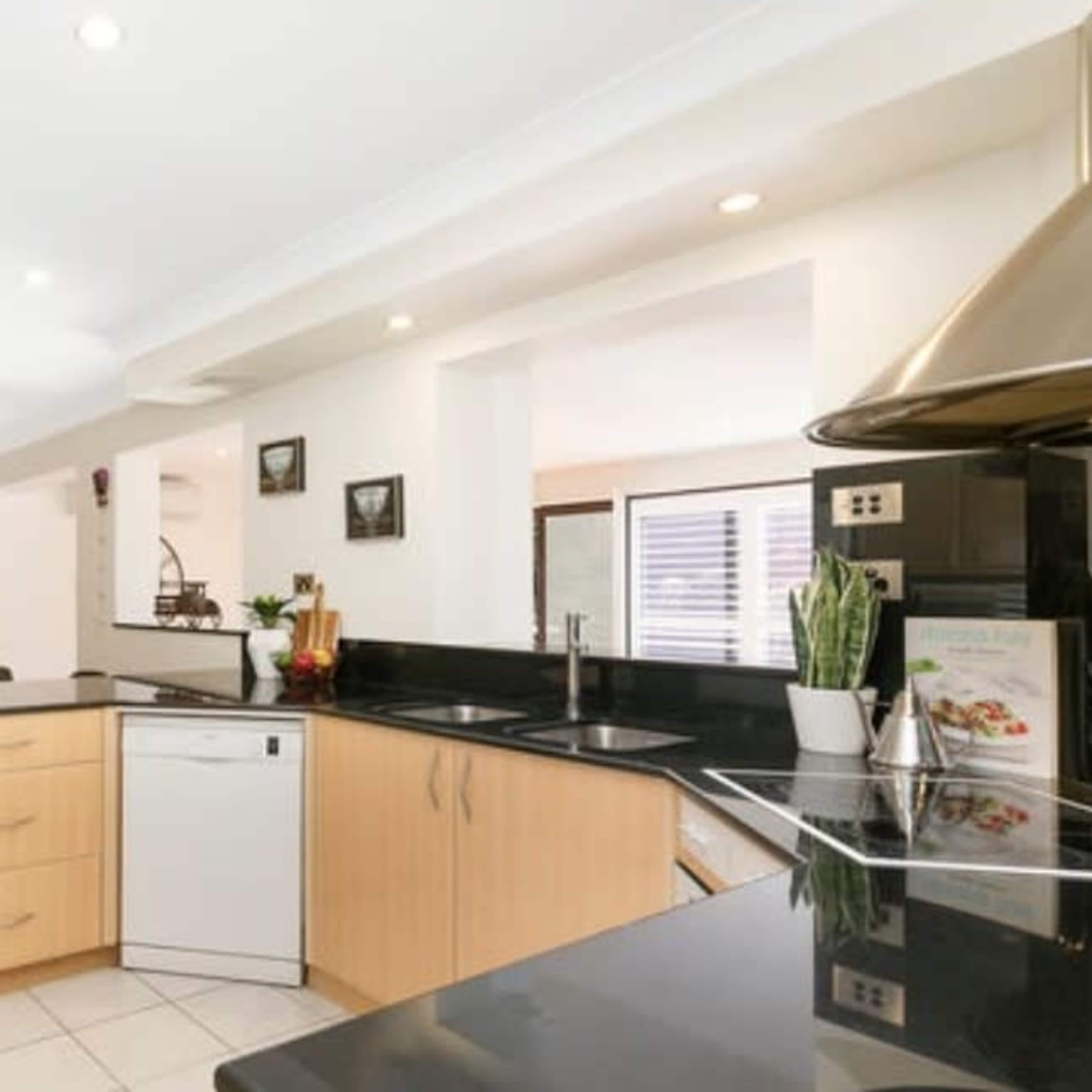 A modern kitchen features light wood cabinetry, black countertops, and stainless steel appliances, with a view into a living area in the background.