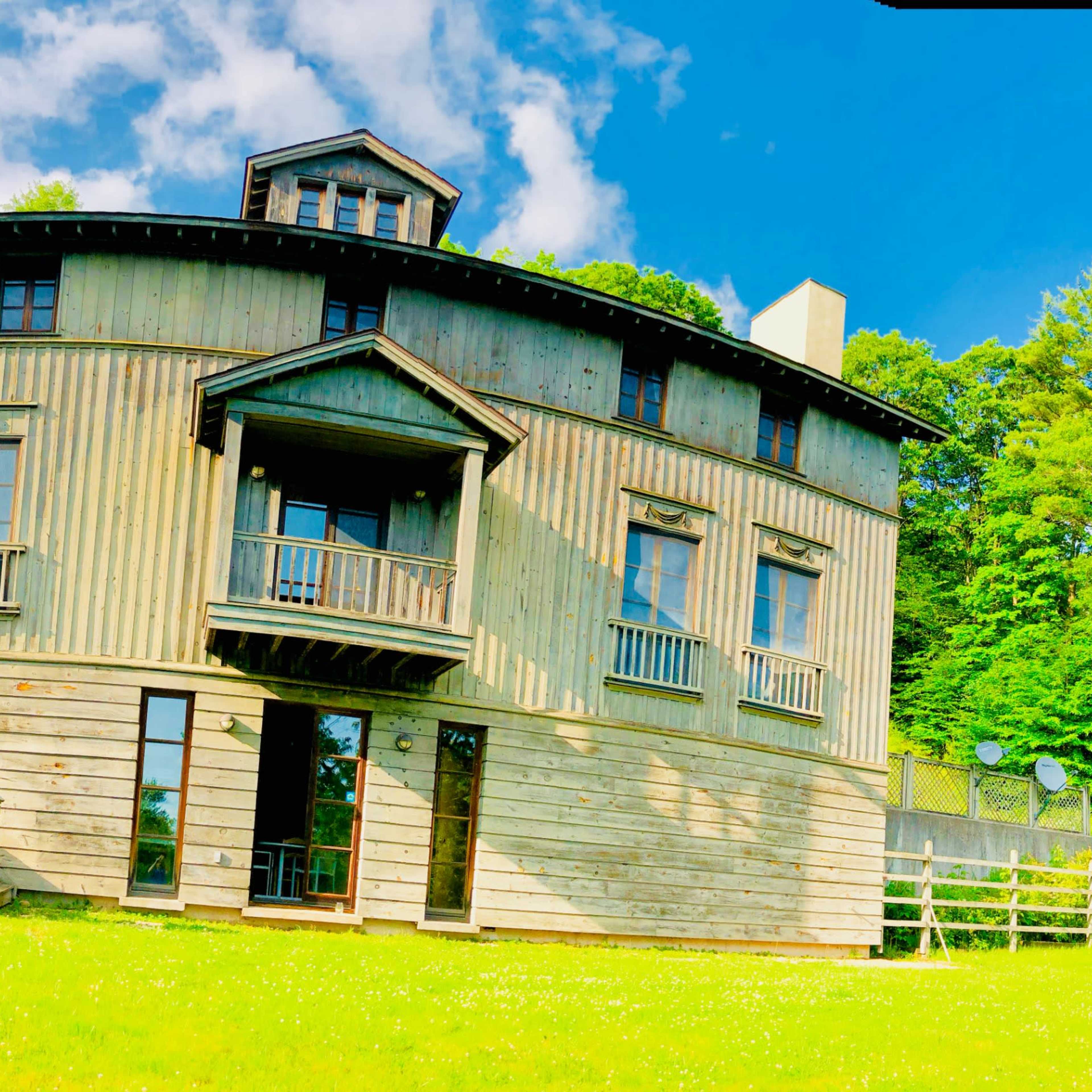 A two-story, wooden house with a unique round design, large windows, and a sloped lawn in front, surrounded by trees and a blue sky.