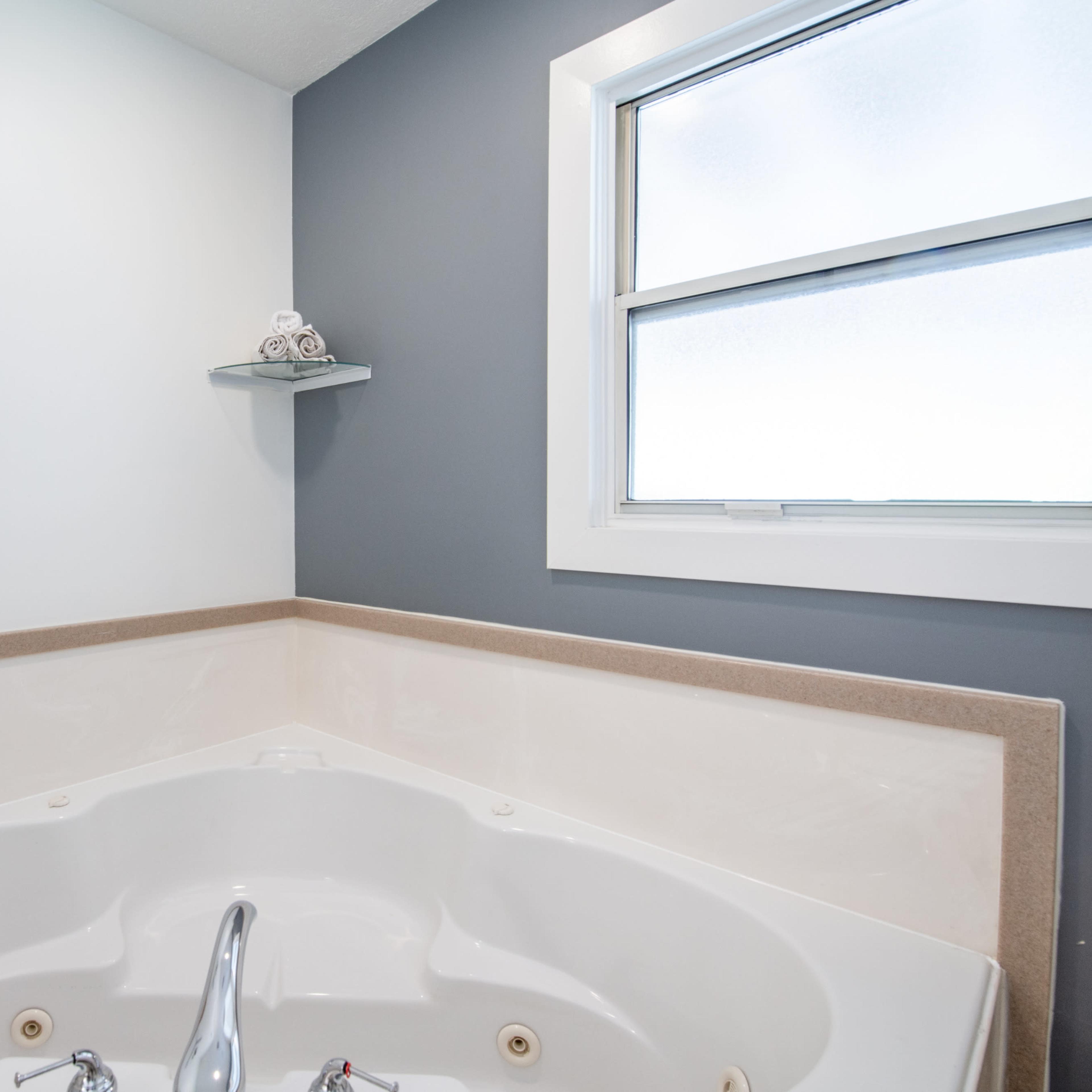 A corner bathtub with a faucet, surrounded by two walls painted in light and dark colors, and a window above it.