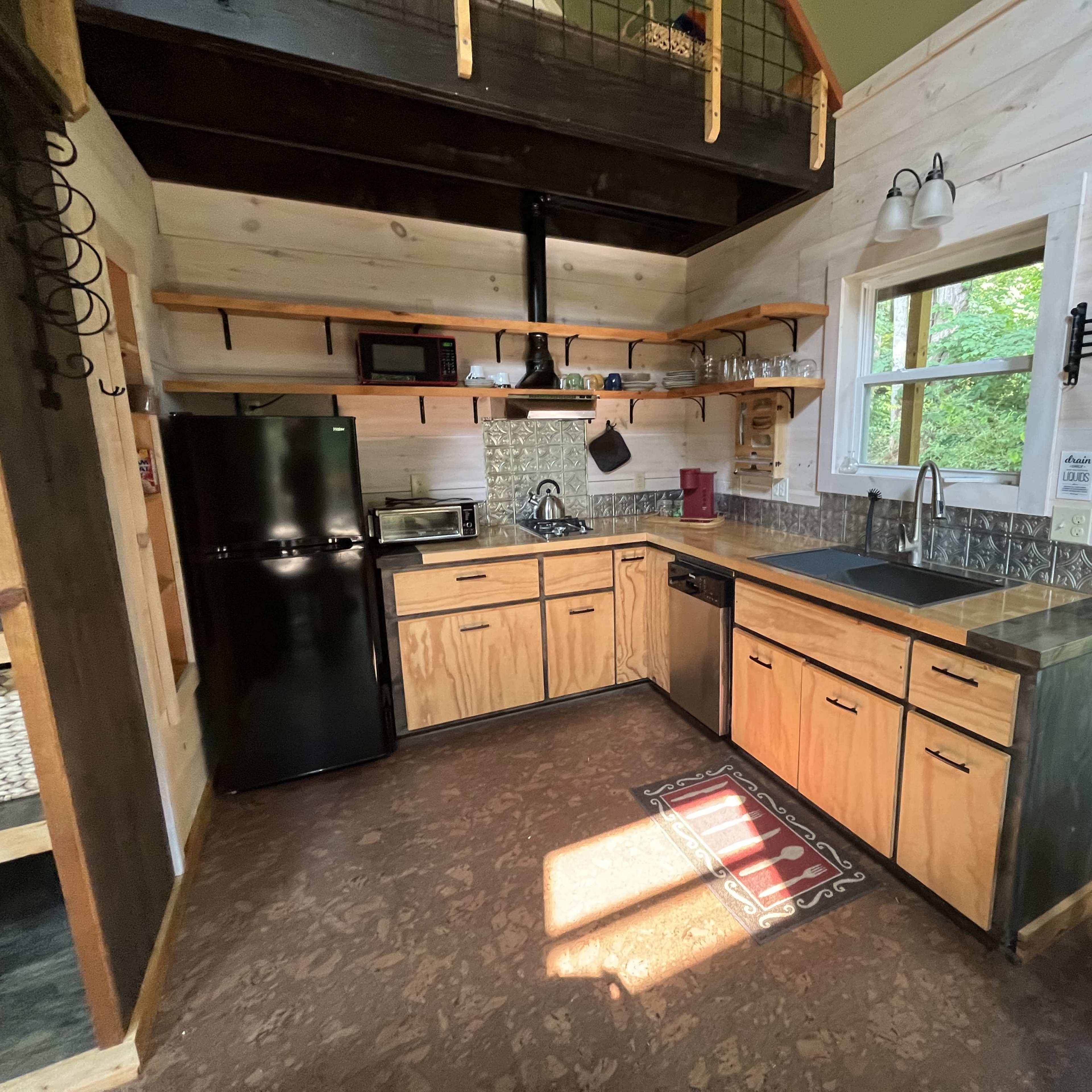 A small kitchen in a wooden cabin featuring a black refrigerator, a microwave, a sink, cabinetry, and a window that provides natural light.