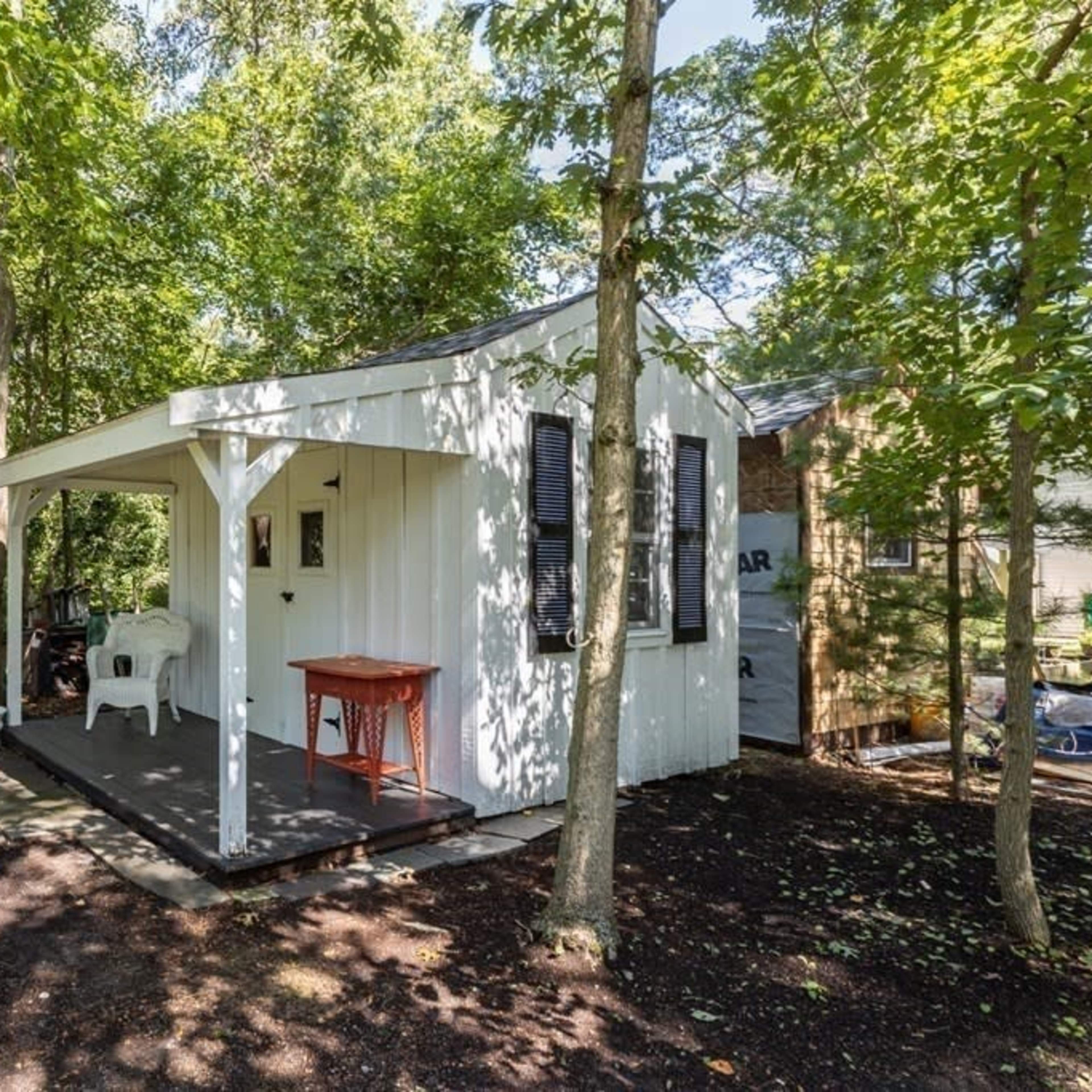A small white cottage with a red table on the porch is nestled among trees in a wooded area.