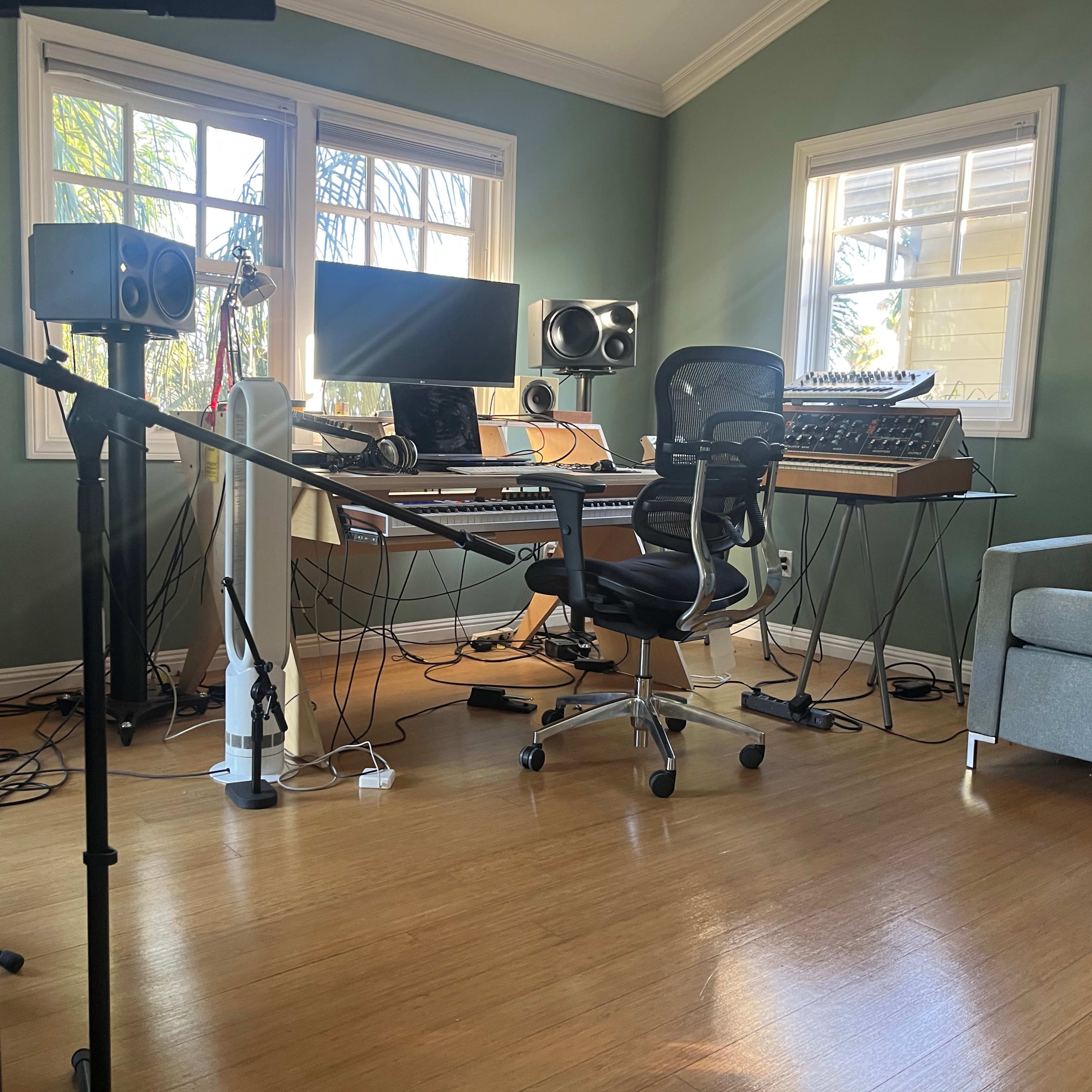 A music production studio features a desk with a computer, keyboards, speakers, and a comfortable chair, surrounded by natural light from the windows.