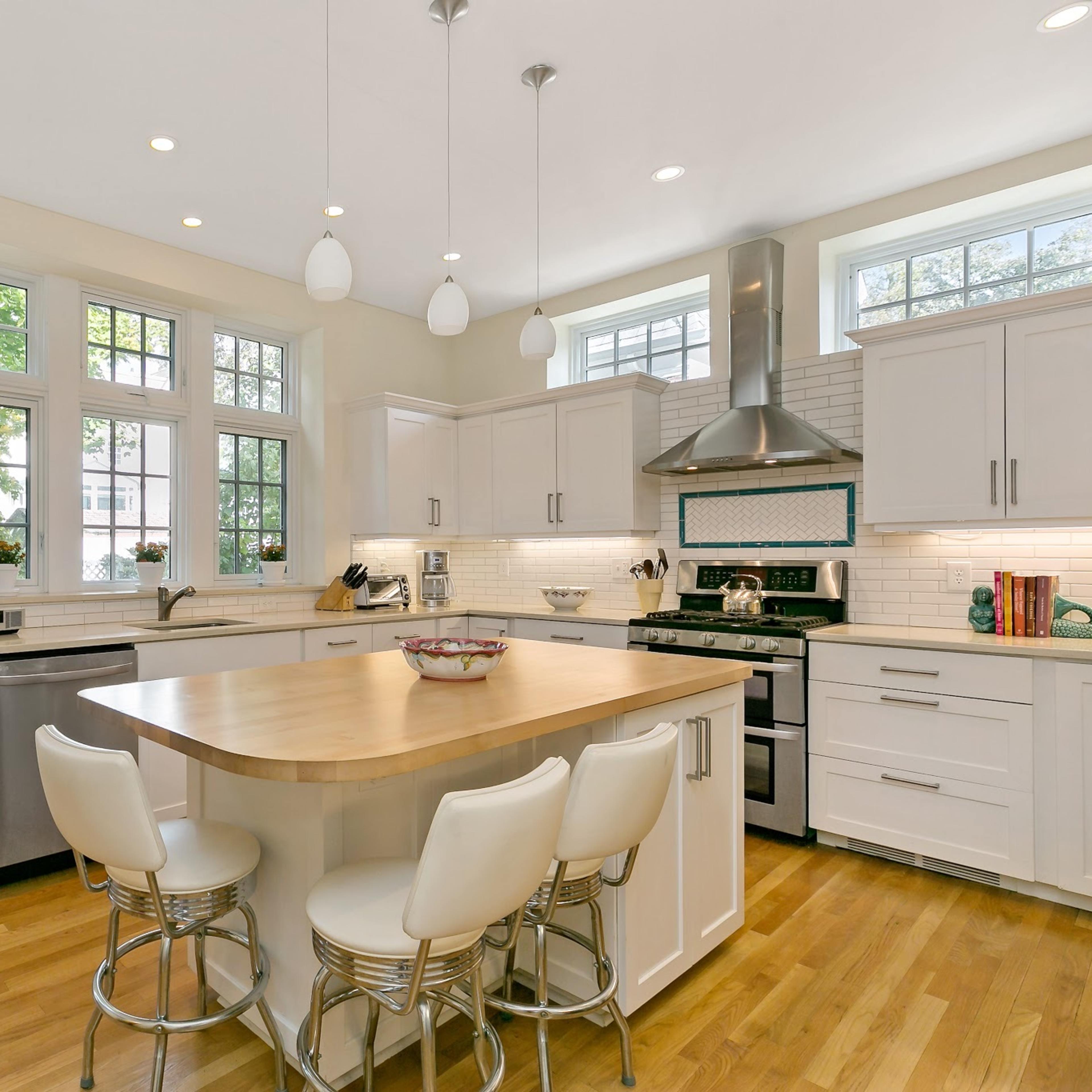 A bright and modern kitchen features white cabinets, a central wooden island with bar stools, and large windows that let in natural light.