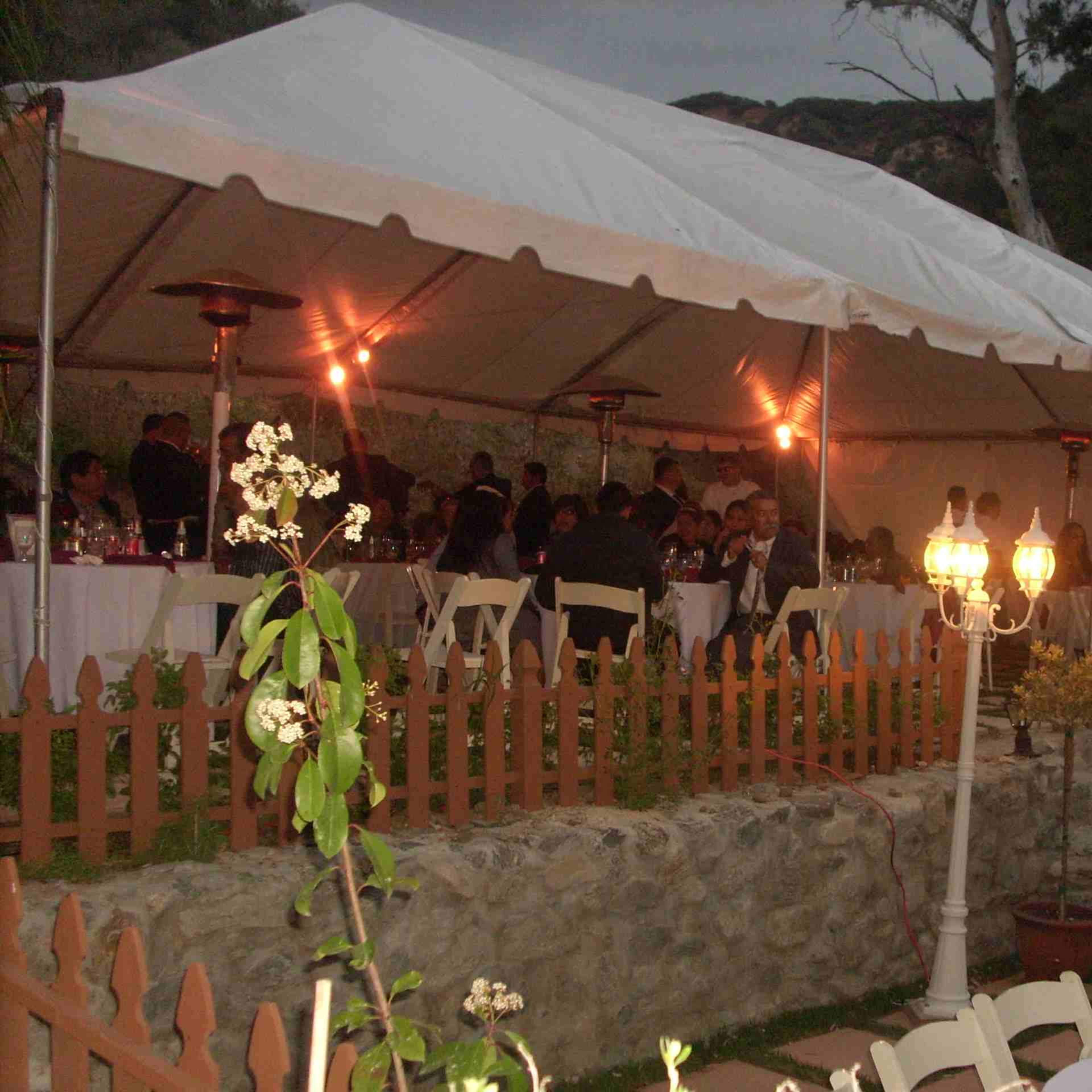 A large tent filled with guests dining at tables is set up in a garden, with outdoor heaters and decorative lighting.