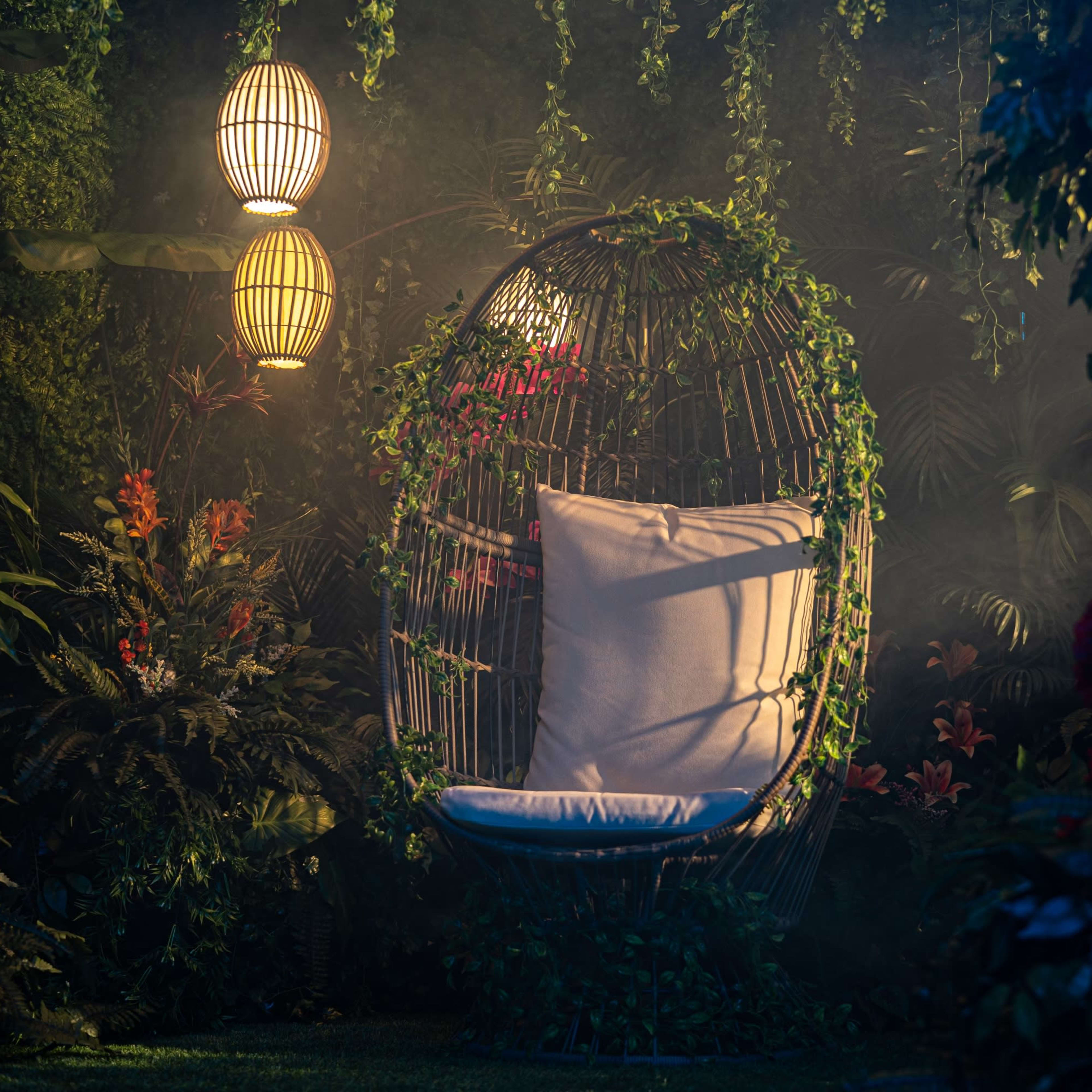 A suspended, wicker egg chair with white cushions is surrounded by lush greenery and illuminated by ambient lanterns.