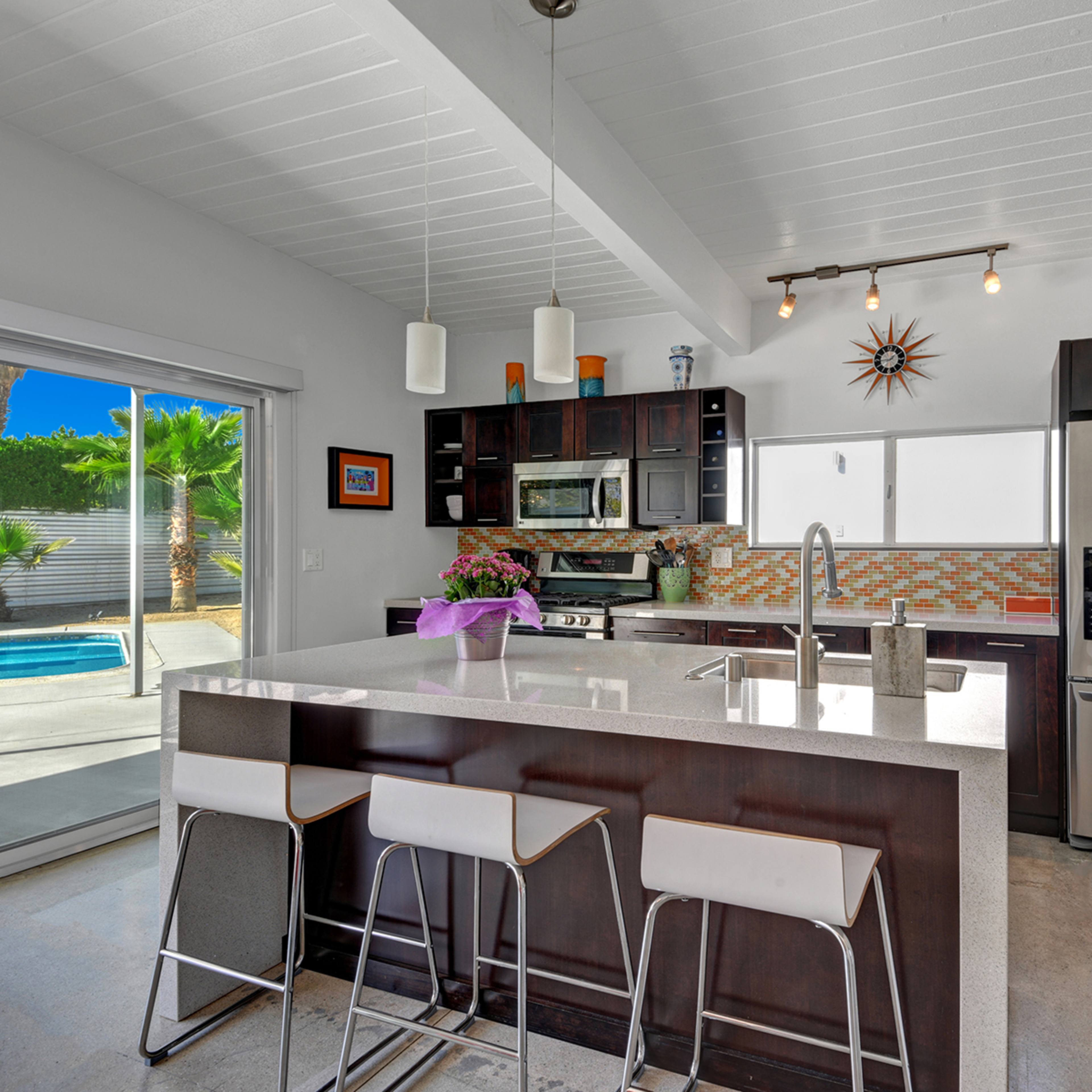 The kitchen features dark wood cabinetry, stainless steel appliances, and a central island with seating, overlooking a patio with a swimming pool through large sliding glass doors.