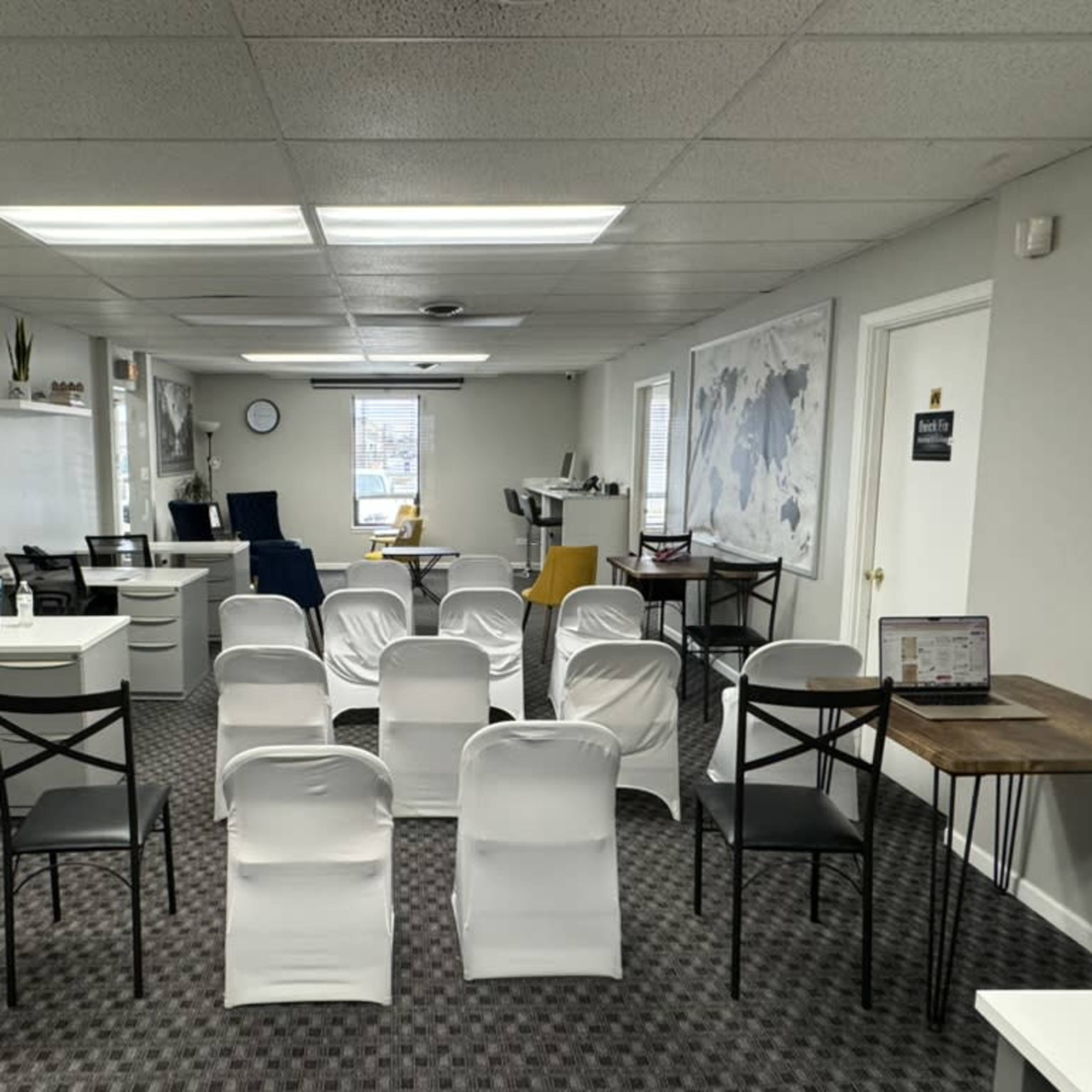 A minimalist office space is arranged with white chairs in rows, flanked by desks and computers, and featuring large windows and a wall map.