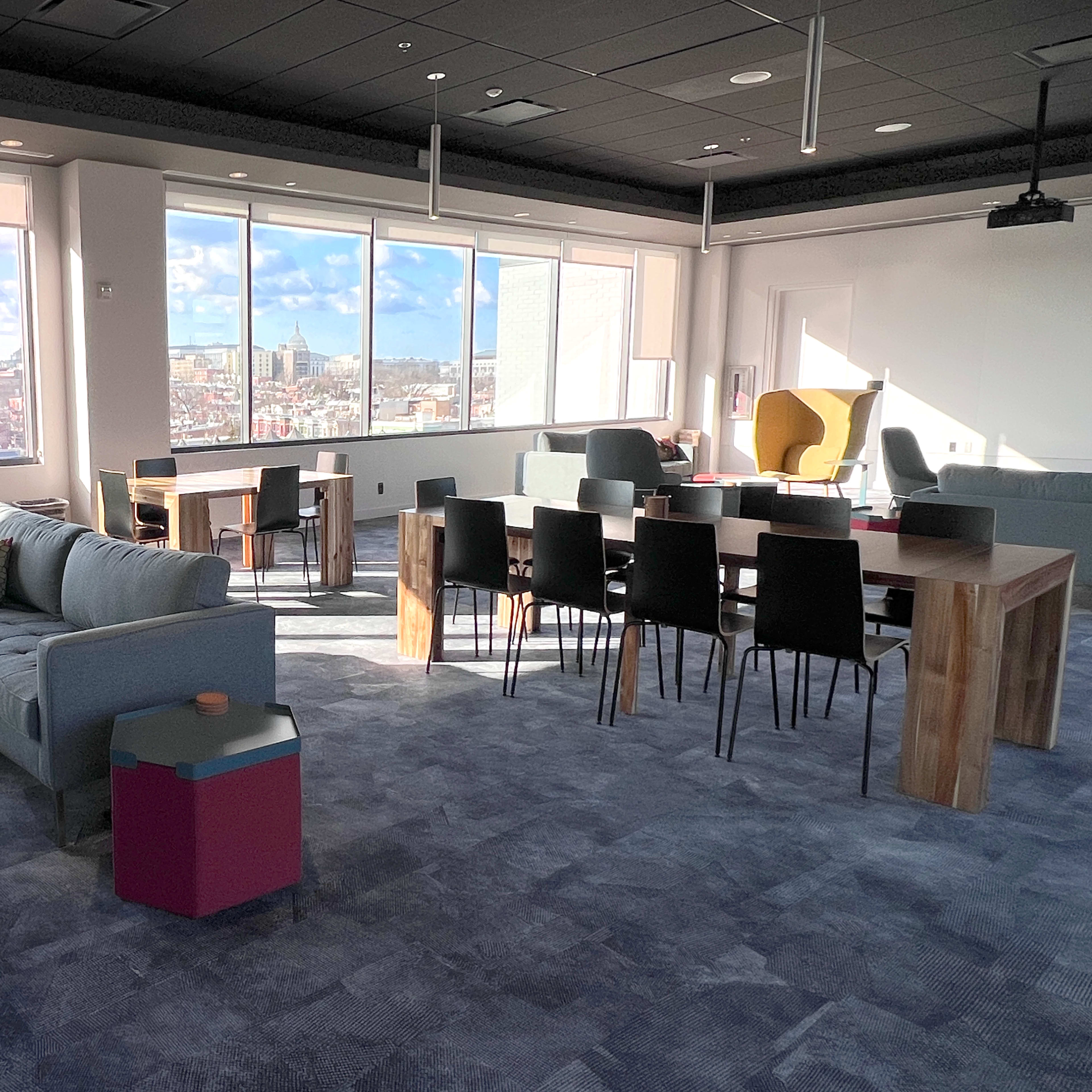 A modern office space featuring a seating area with a couch and chairs, a dining table, and large windows overlooking a city skyline.