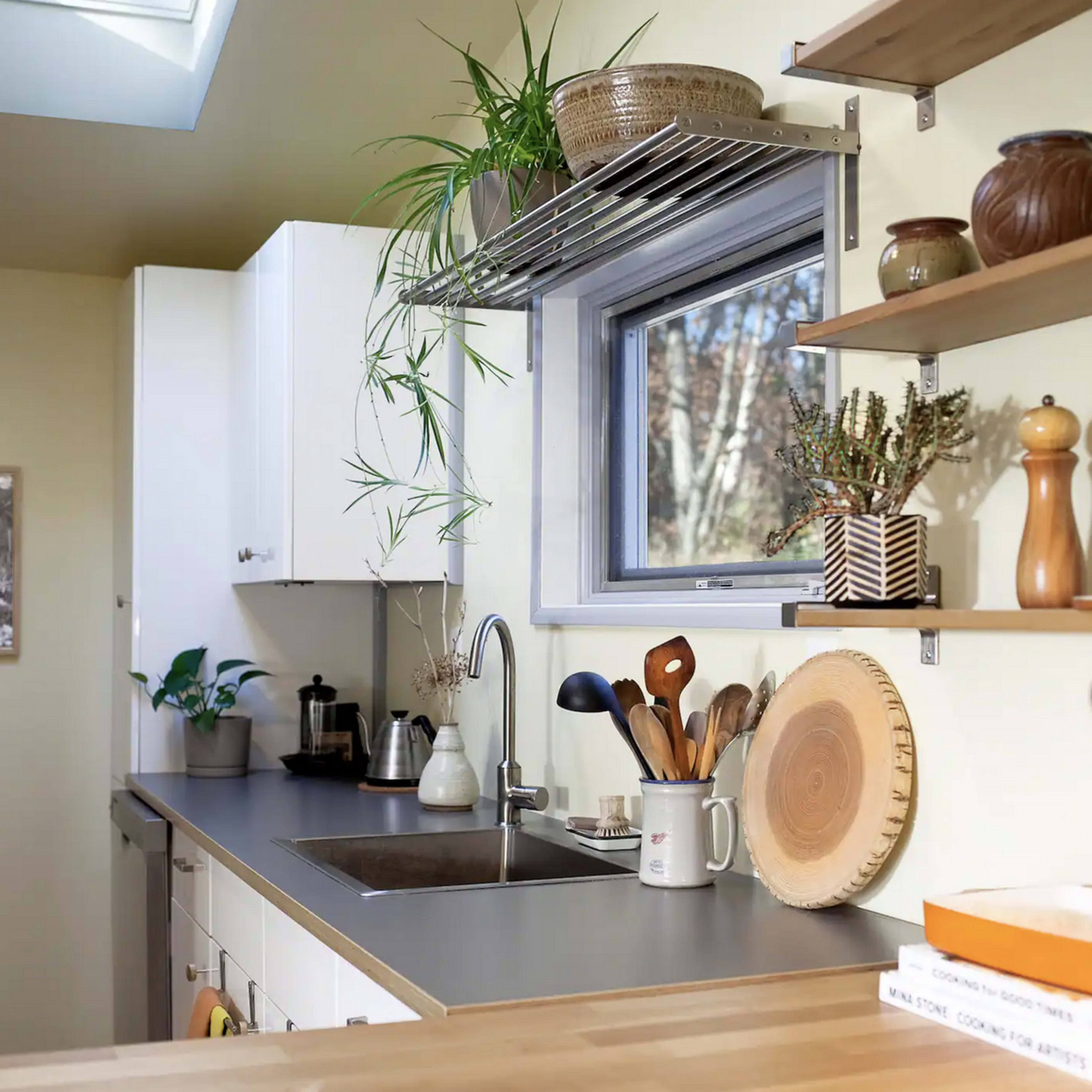 A modern kitchen features a countertop with a bowl of fruit, shelves with kitchenware, and a sink under a window with natural light.