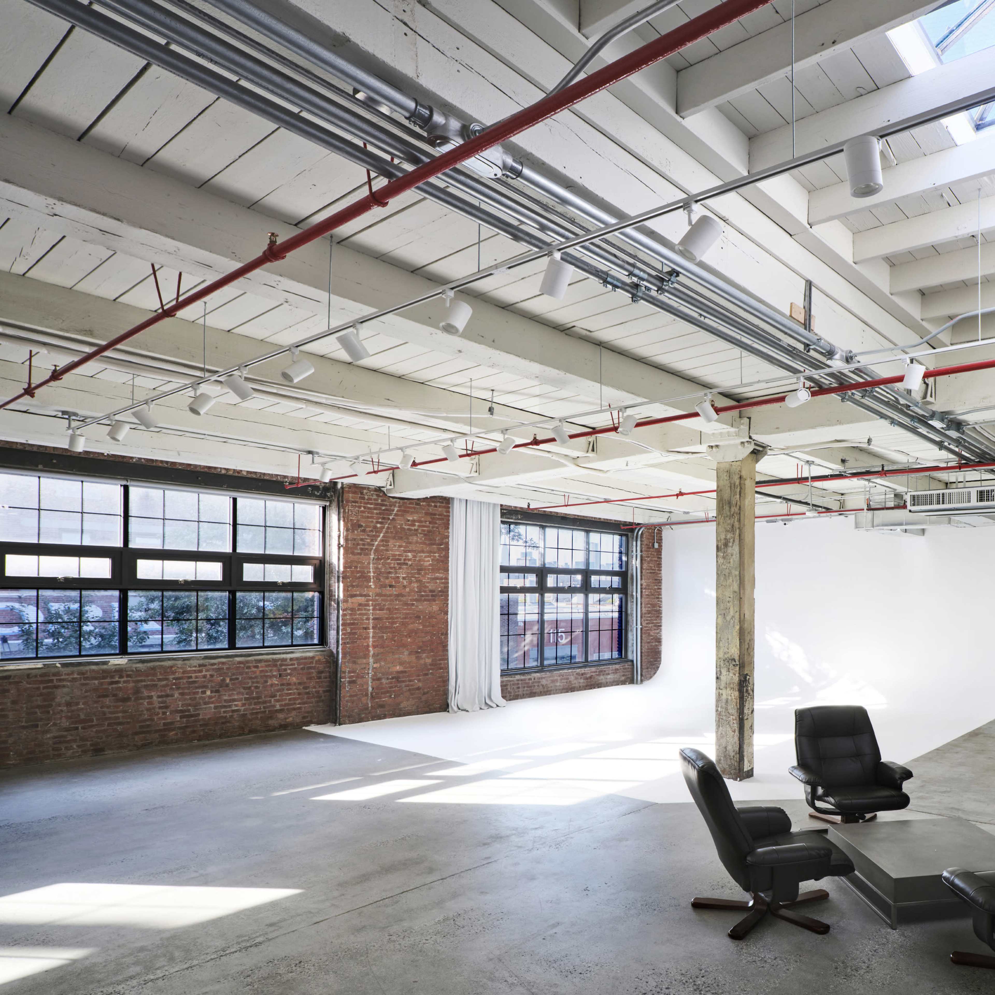 A spacious, modern interior with exposed brick walls, a concrete floor, and large windows allowing natural light to enter a minimalist meeting area featuring black chairs and a small table.