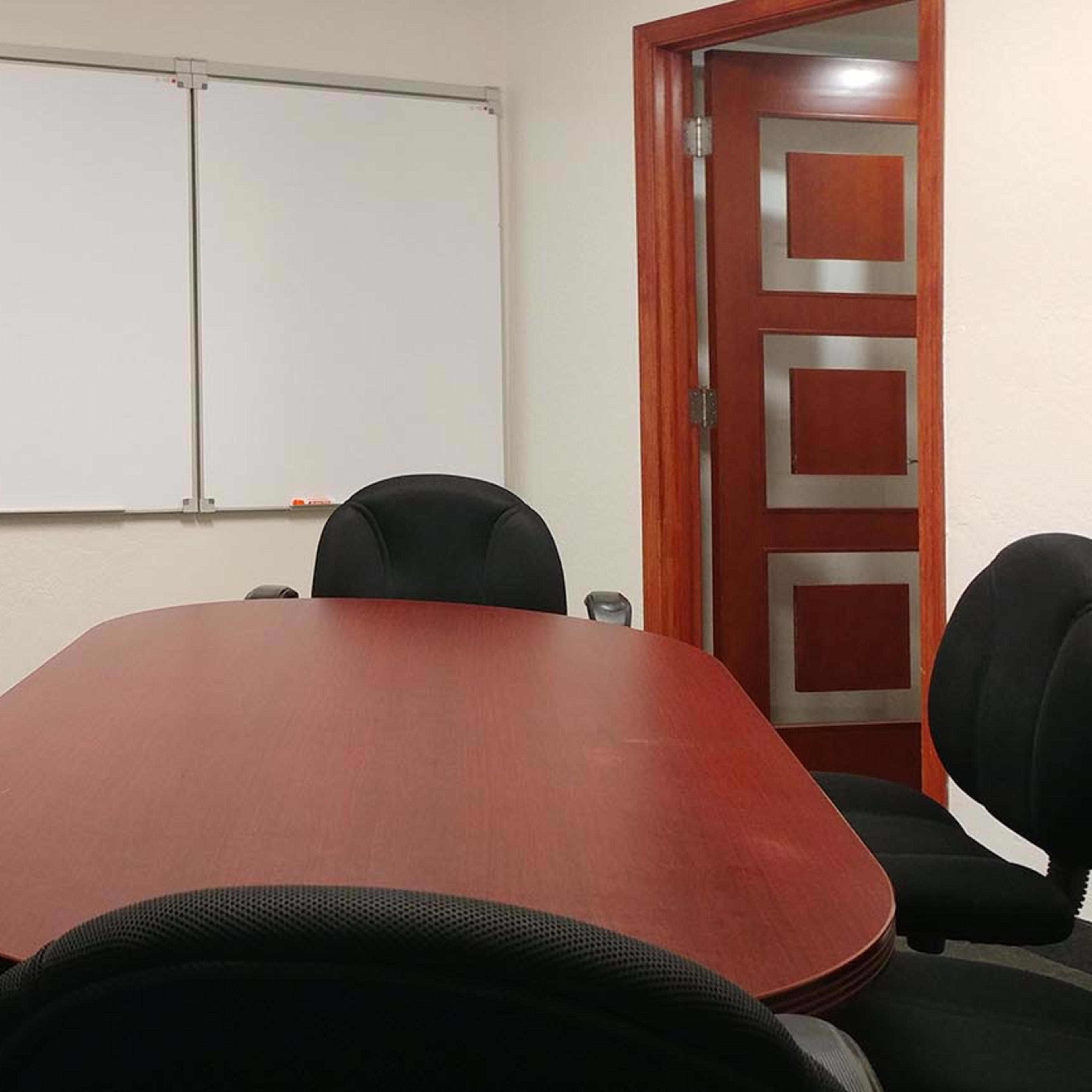 A conference room with a wooden table surrounded by black chairs and empty whiteboards on the wall.