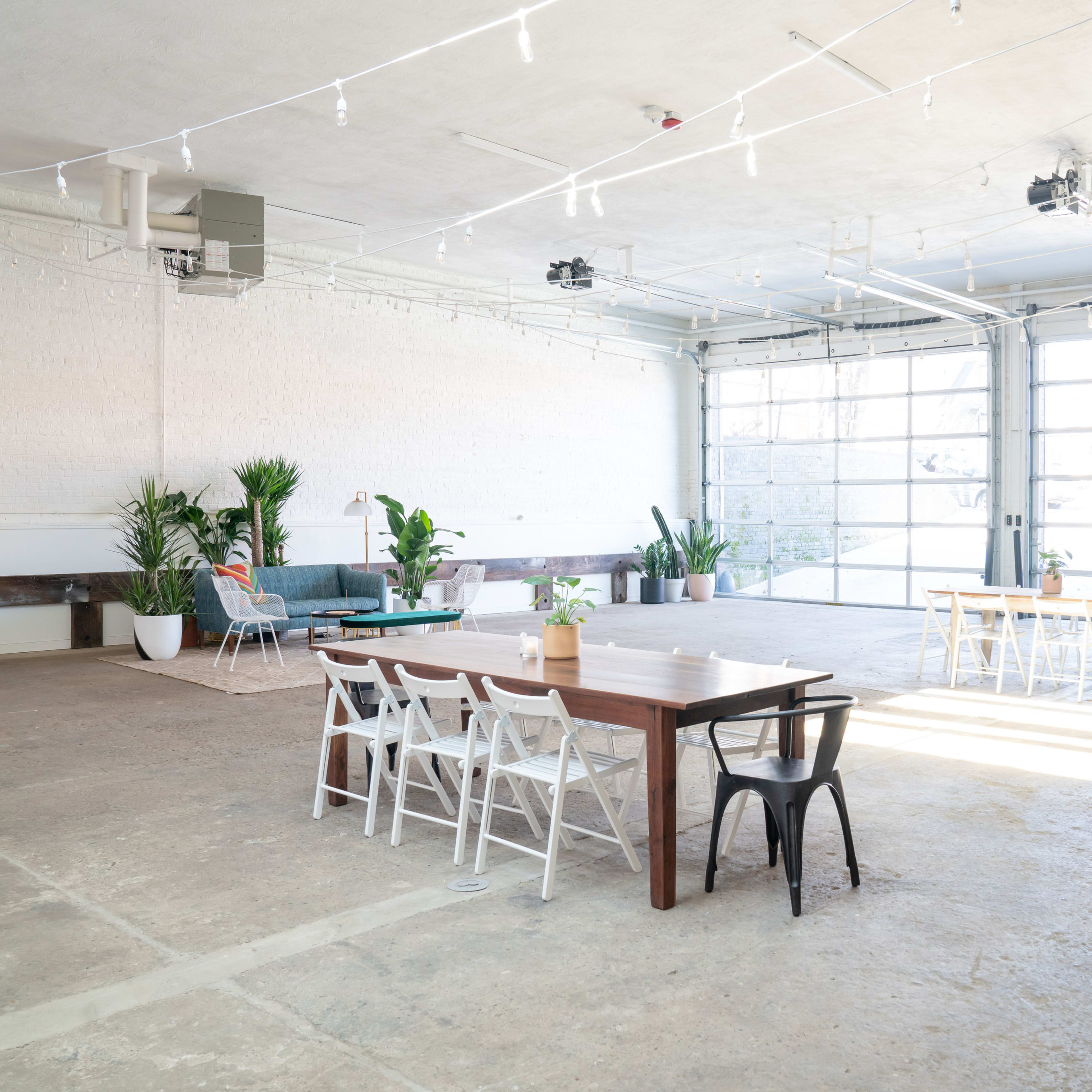 A spacious, well-lit industrial room with large windows, featuring two tables surrounded by chairs and various indoor plants.