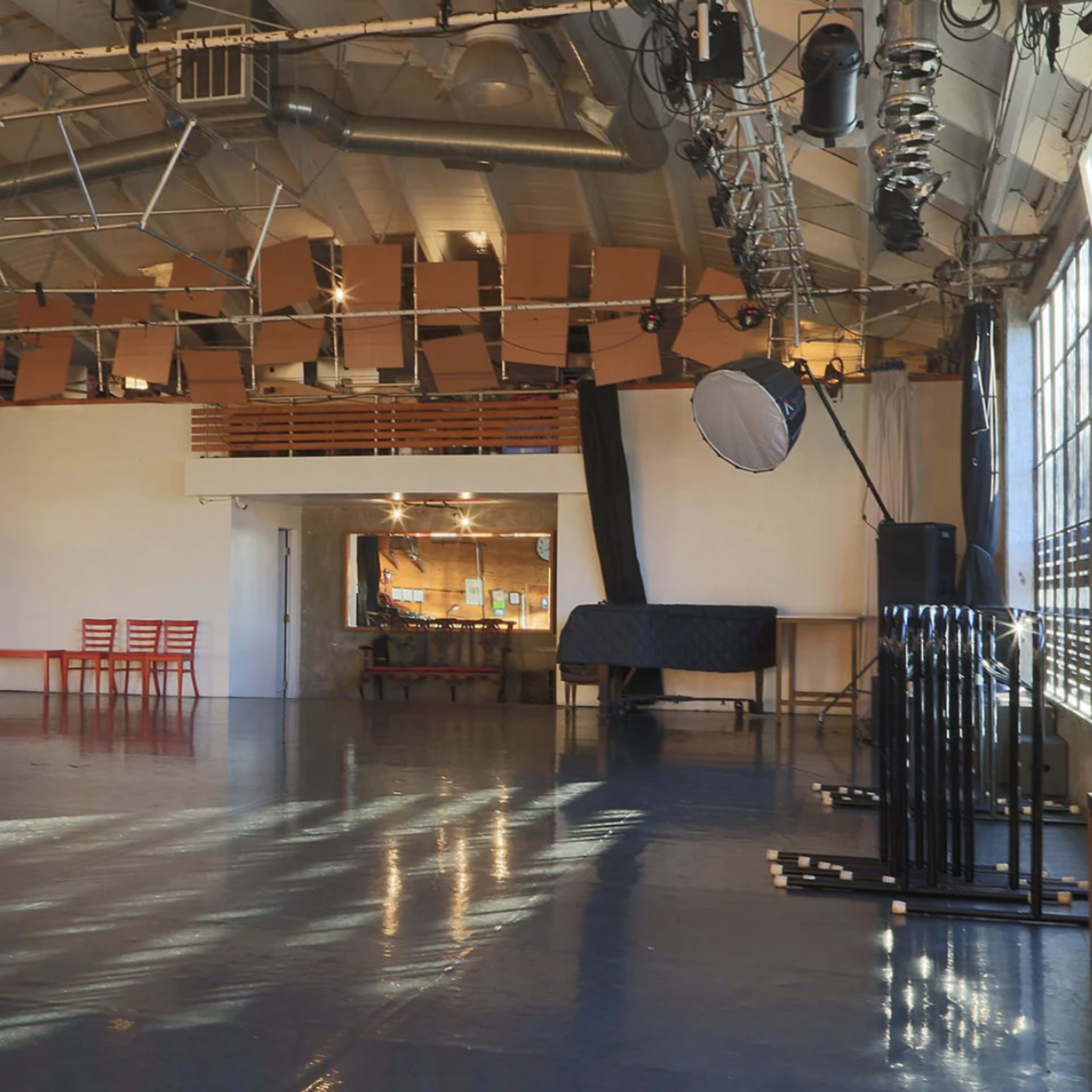 A spacious, well-lit studio with a polished floor, large windows, and several red chairs arranged near a mirror.