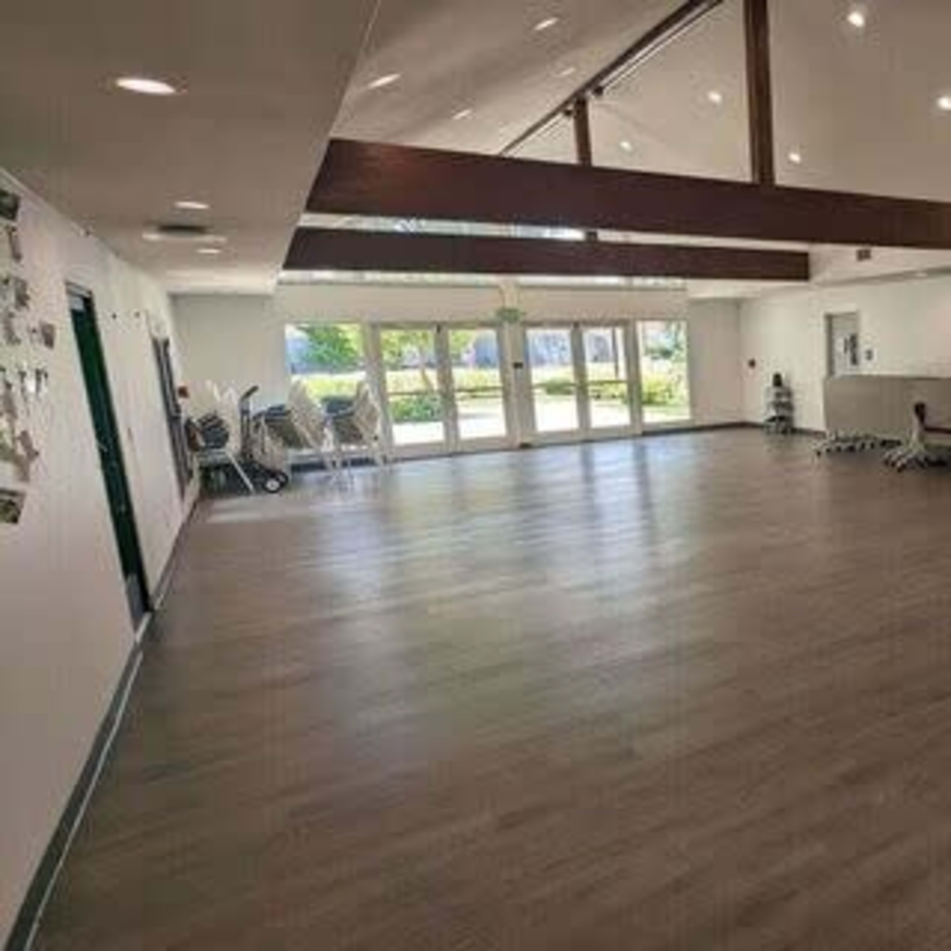 A spacious, brightly lit room with wooden flooring and large windows, featuring exercise equipment along one wall.