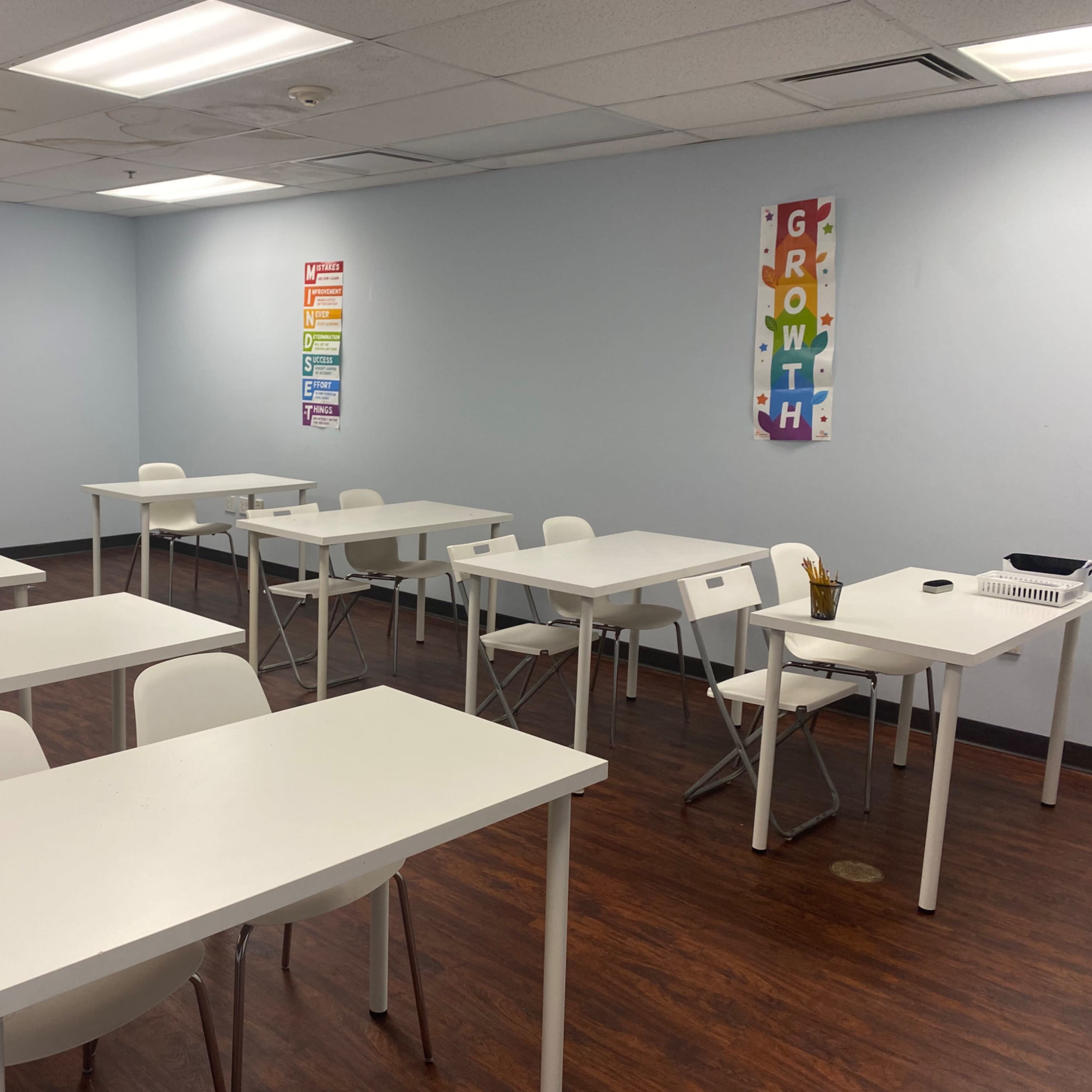 A classroom with white tables and chairs arranged in a sparse layout, featuring posters on the walls.