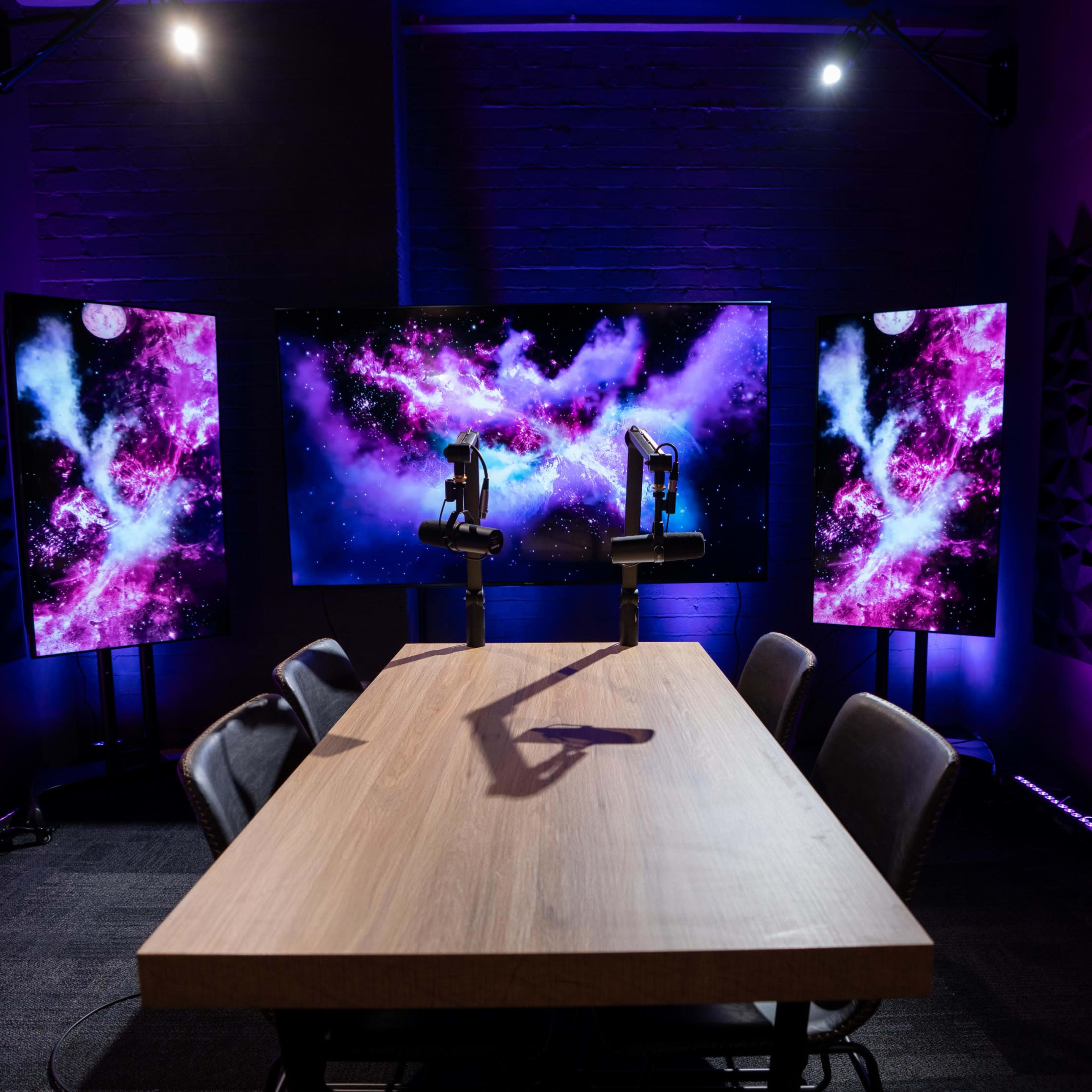A conference room features a wooden table with black chairs, flanked by two large screens displaying a colorful cosmic background.