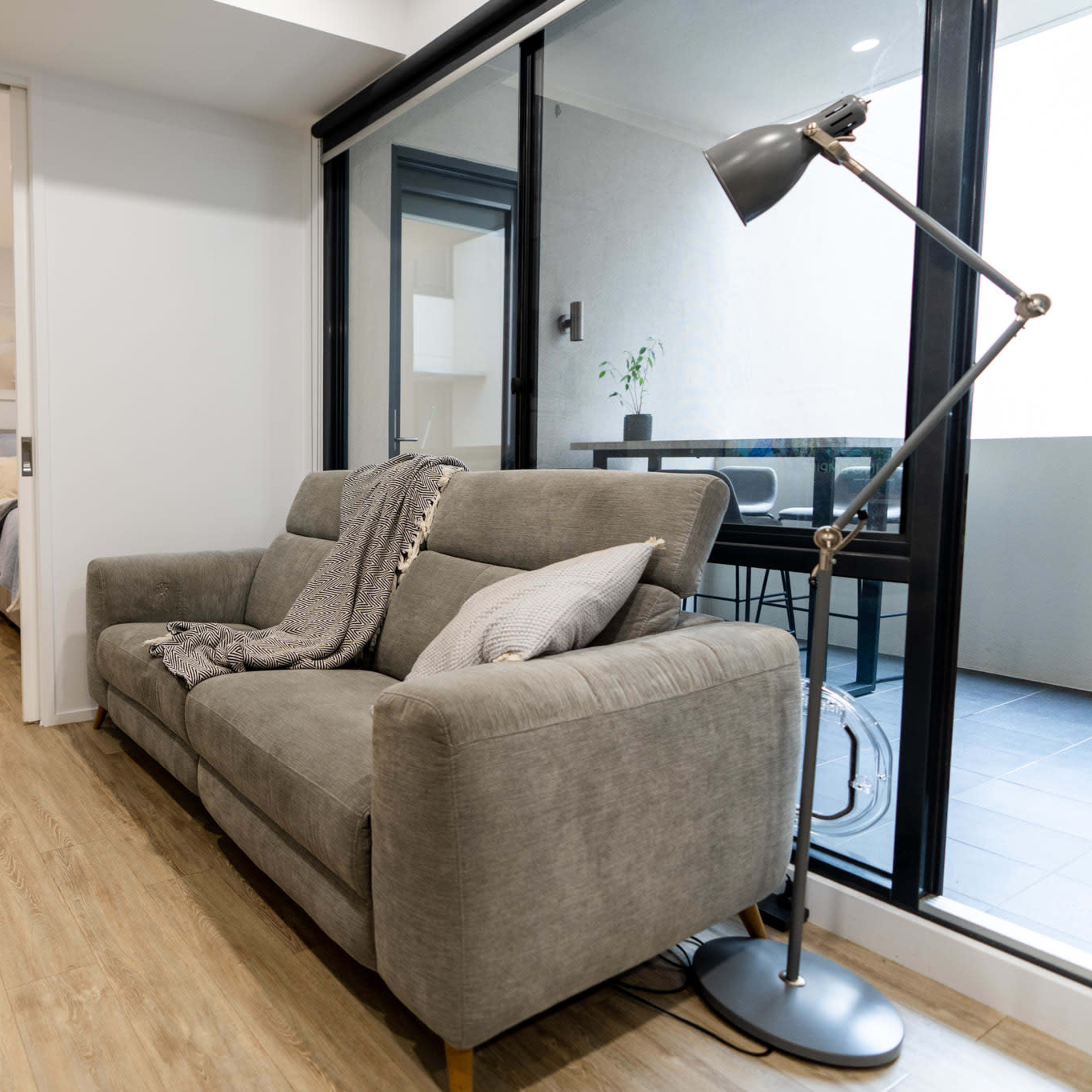 A gray sofa is placed in a modern, open living area with large windows and a view of a balcony.