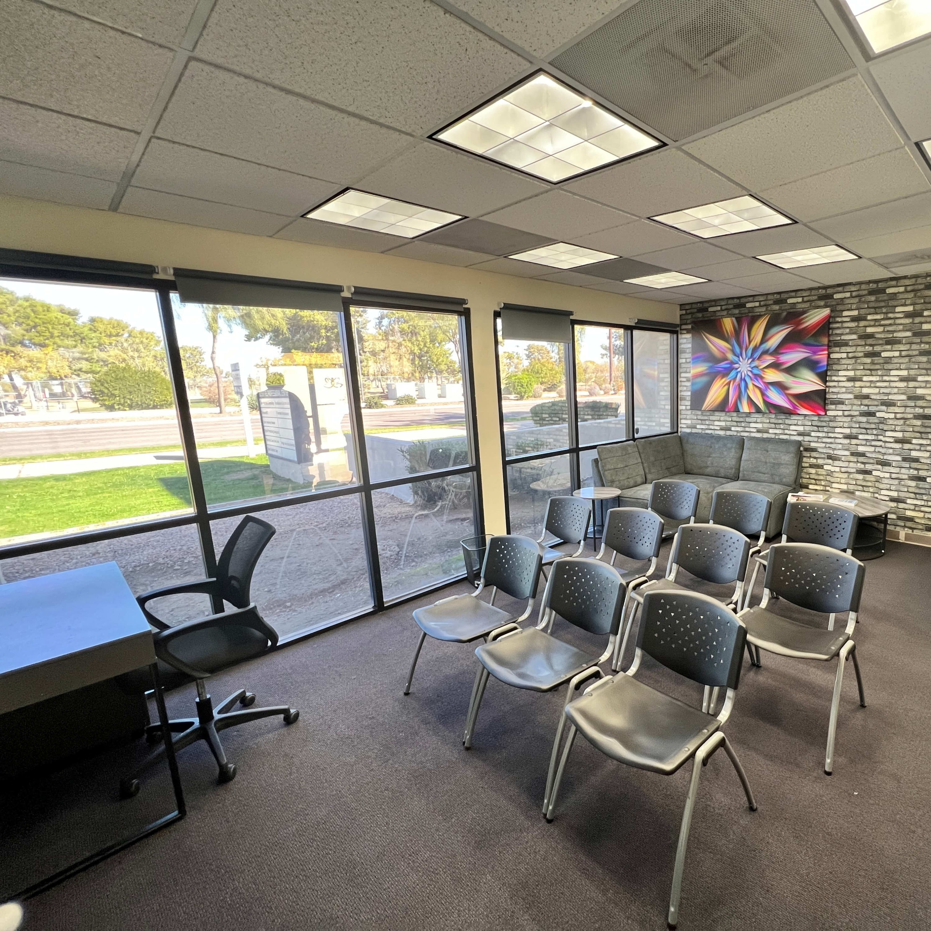A conference room with several gray chairs arranged in front of a wall featuring a colorful artwork, alongside windows that provide outdoor views.