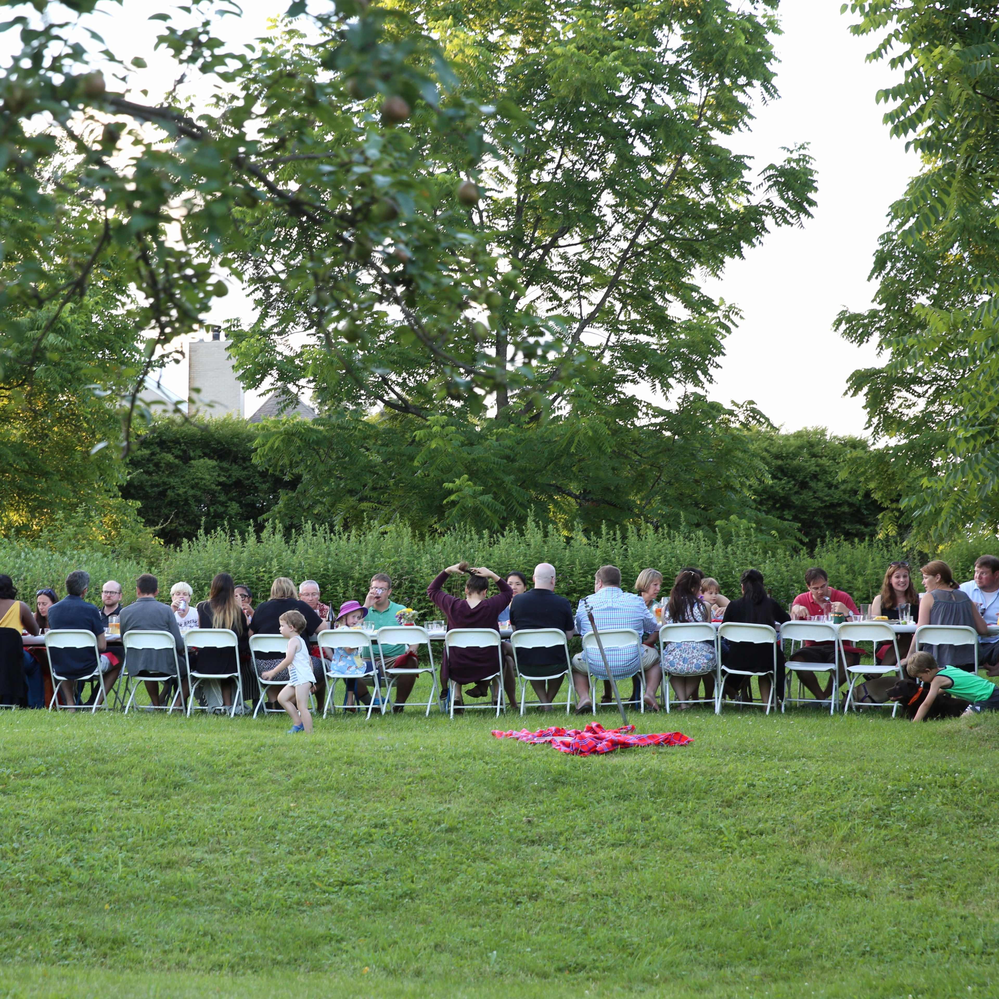 A long table is set outdoors with many people seated around it during a gathering in a grassy area.