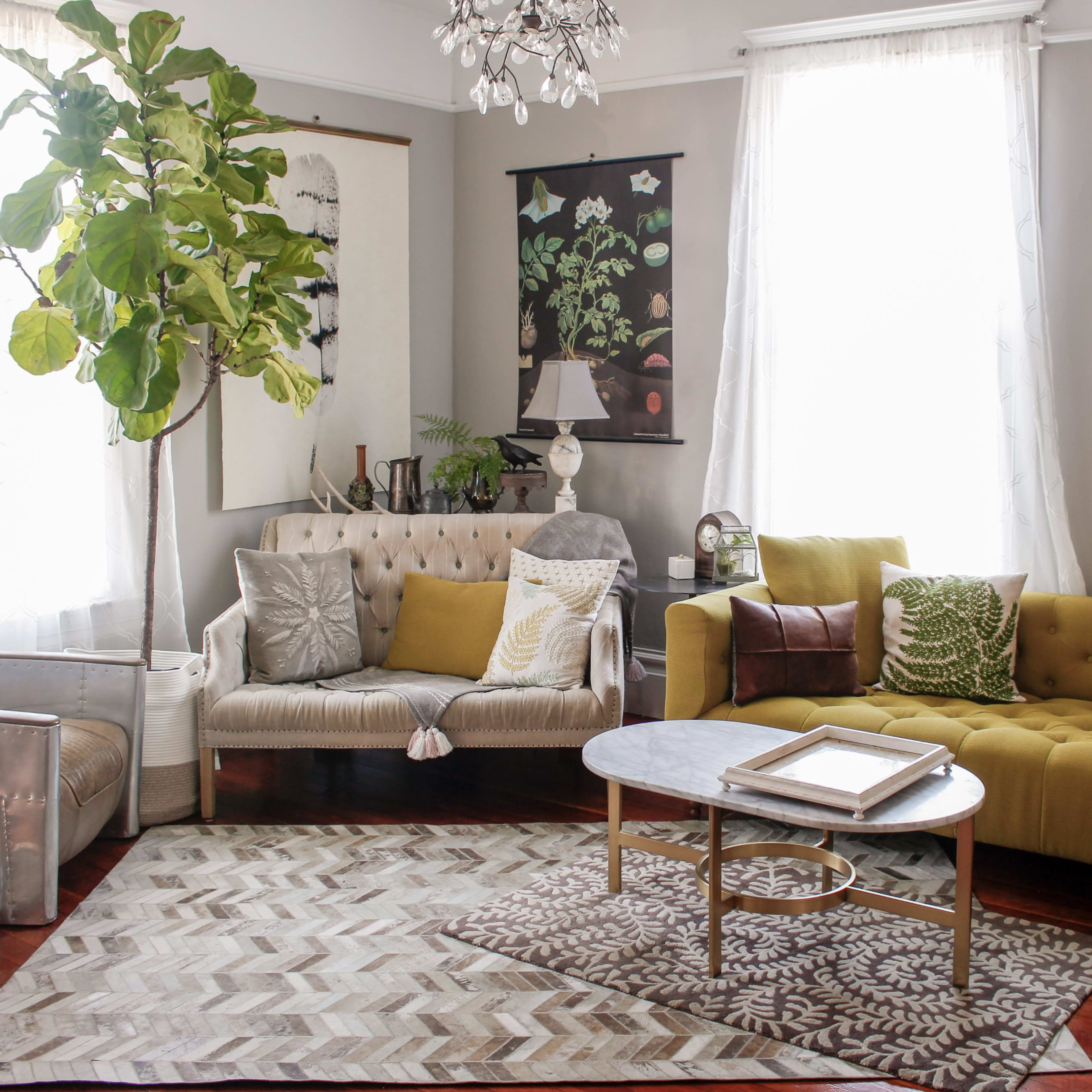 A bright living room features a mix of beige and green sofas, a round coffee table, and plants, with large windows allowing natural light to fill the space.