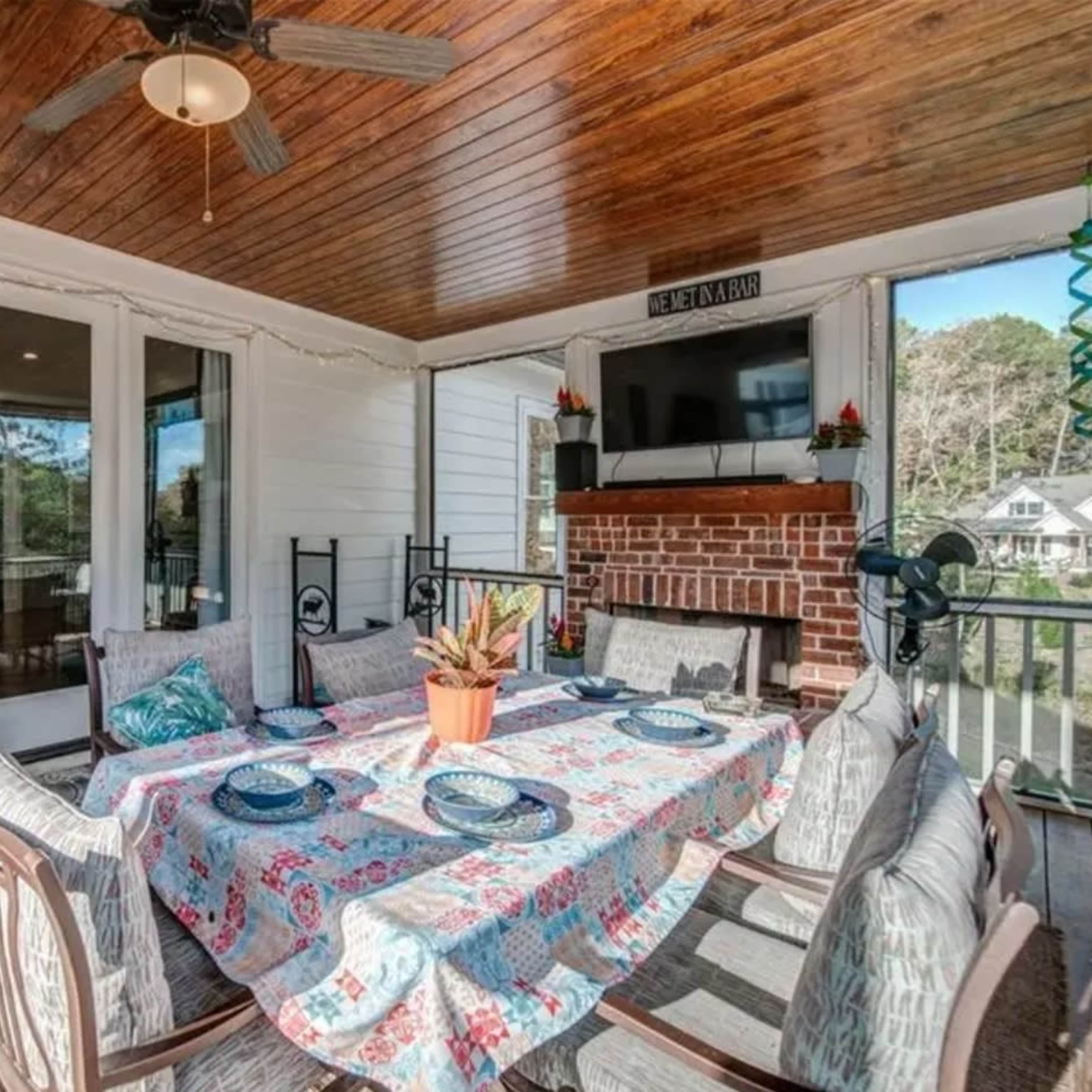 A screened patio features a dining table set with a colorful tablecloth, surrounded by chairs and a brick fireplace, with large windows offering a view of the outdoors.
