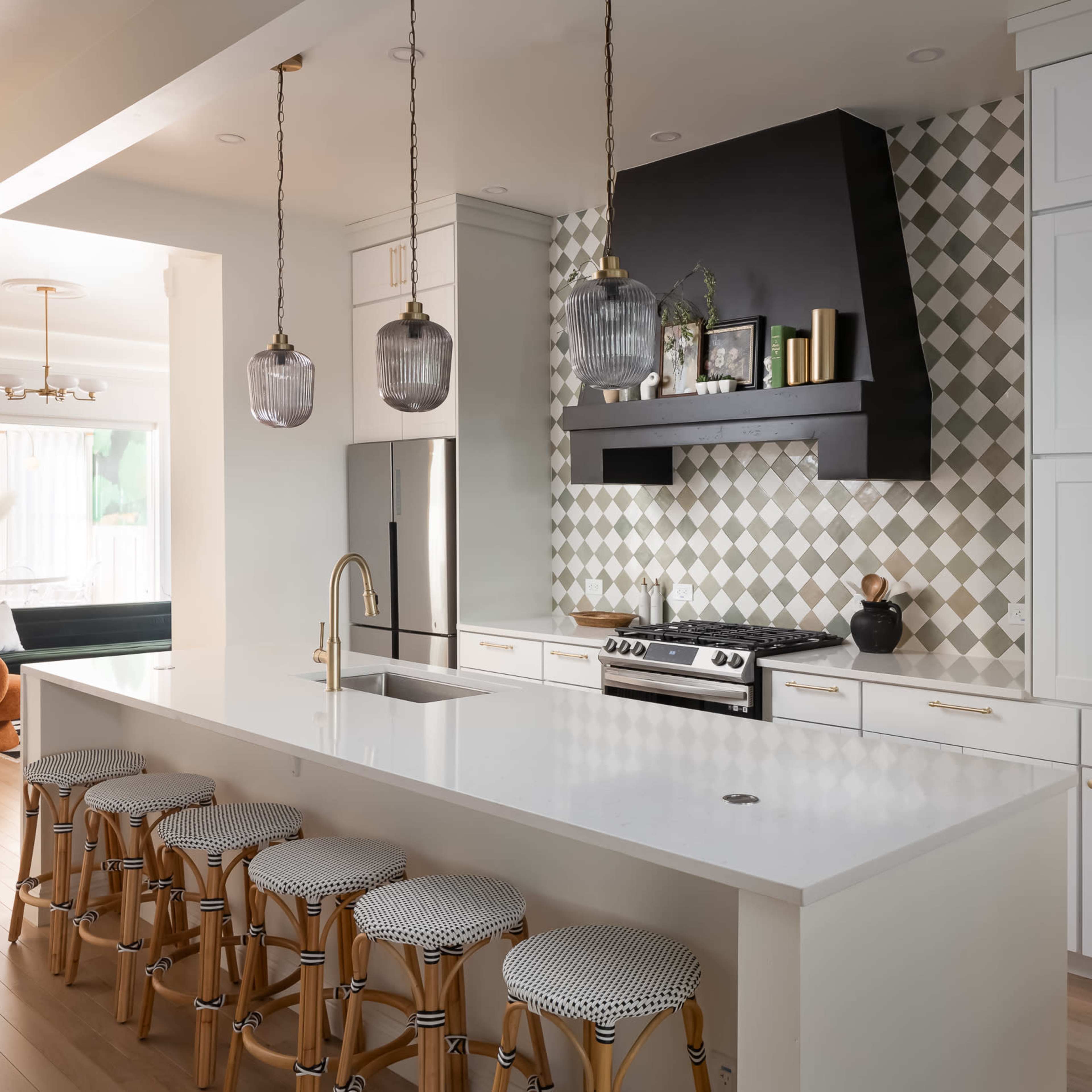 A modern kitchen features a large island with bar stools, stylish pendant lighting, and a patterned backsplash behind the stove.