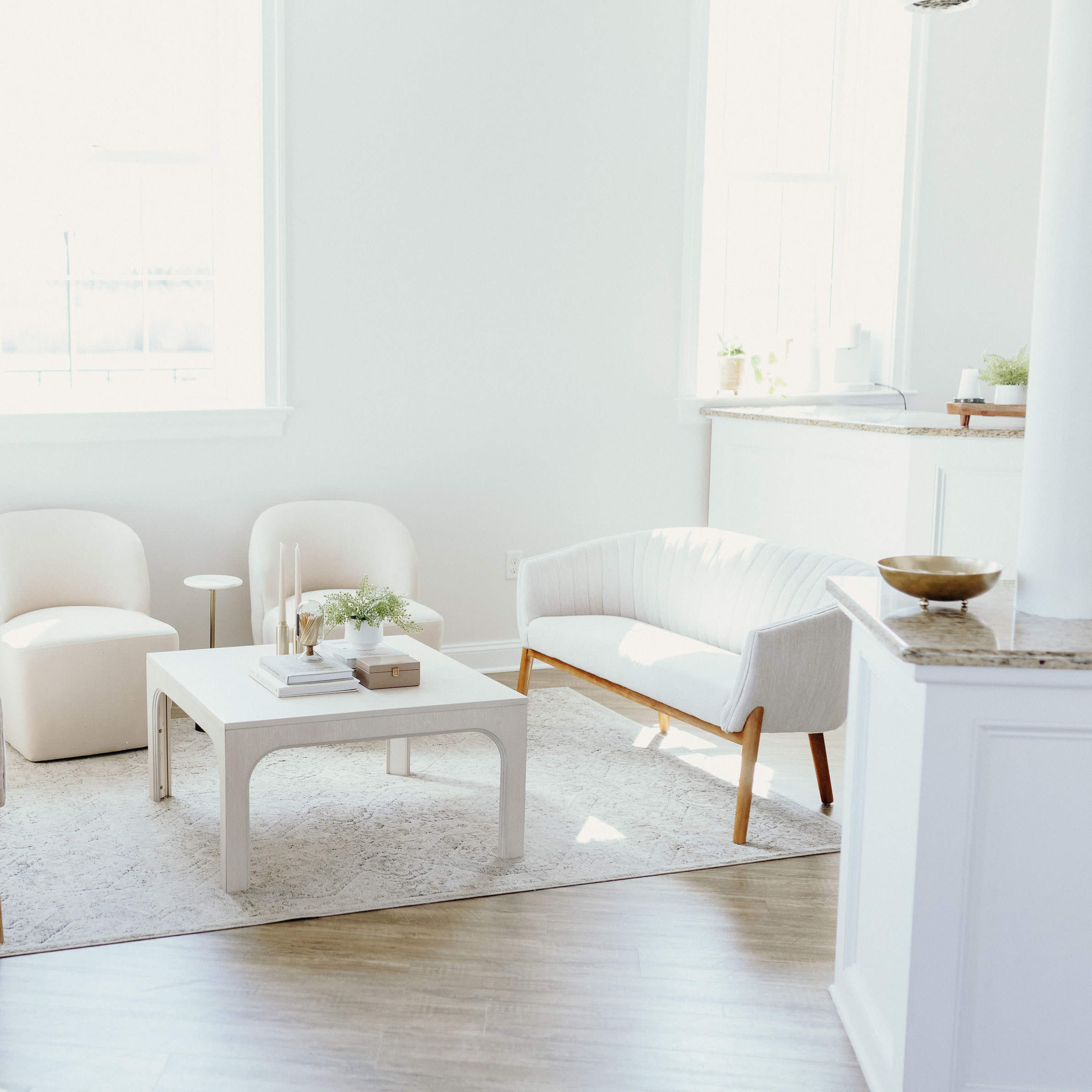 A bright, minimalistic living room with two white armchairs, one light-colored chair, and a square table on a light rug.