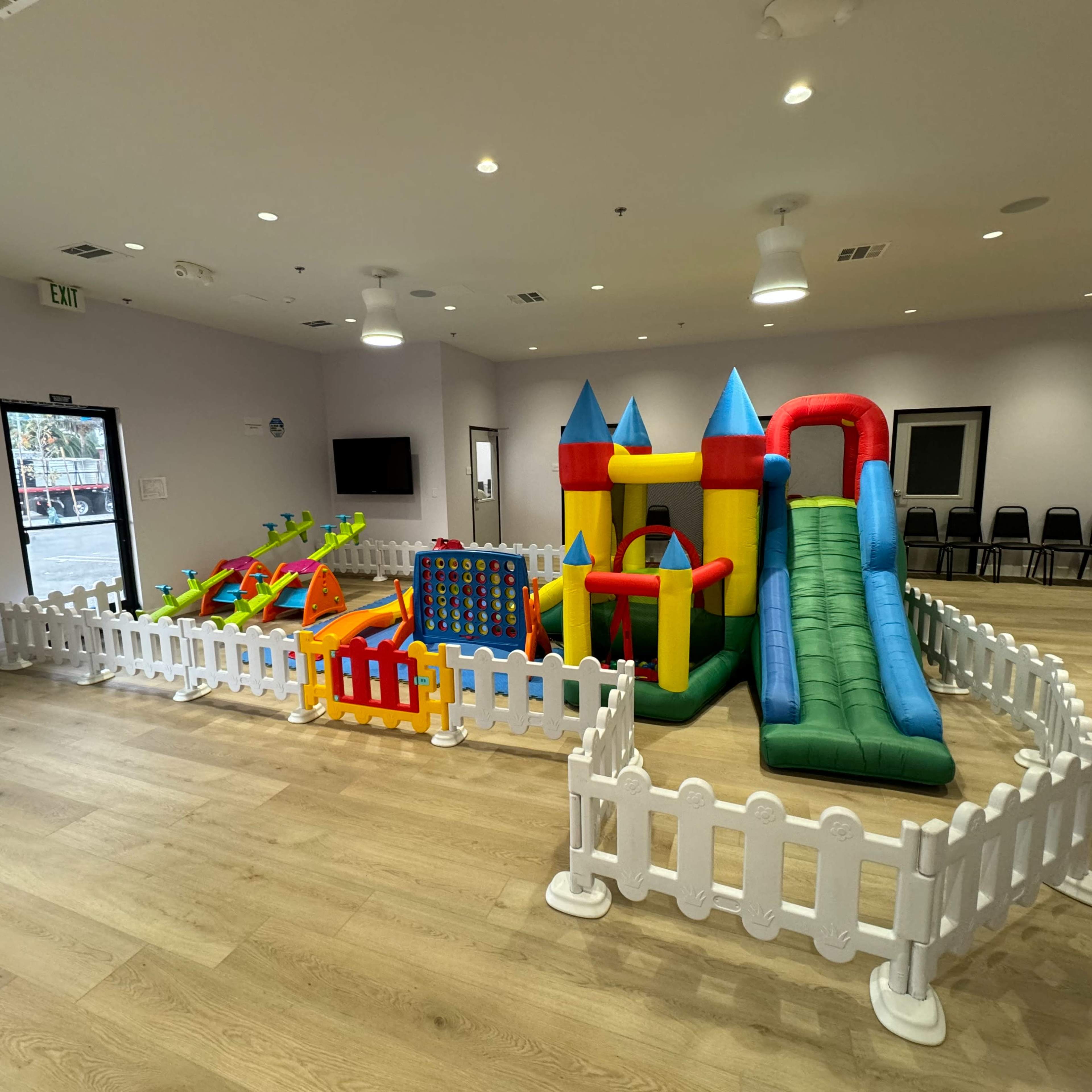 A colorful inflatable play area enclosed by a white safety fence, featuring slides, climbing structures, and various play elements in a well-lit indoor space.