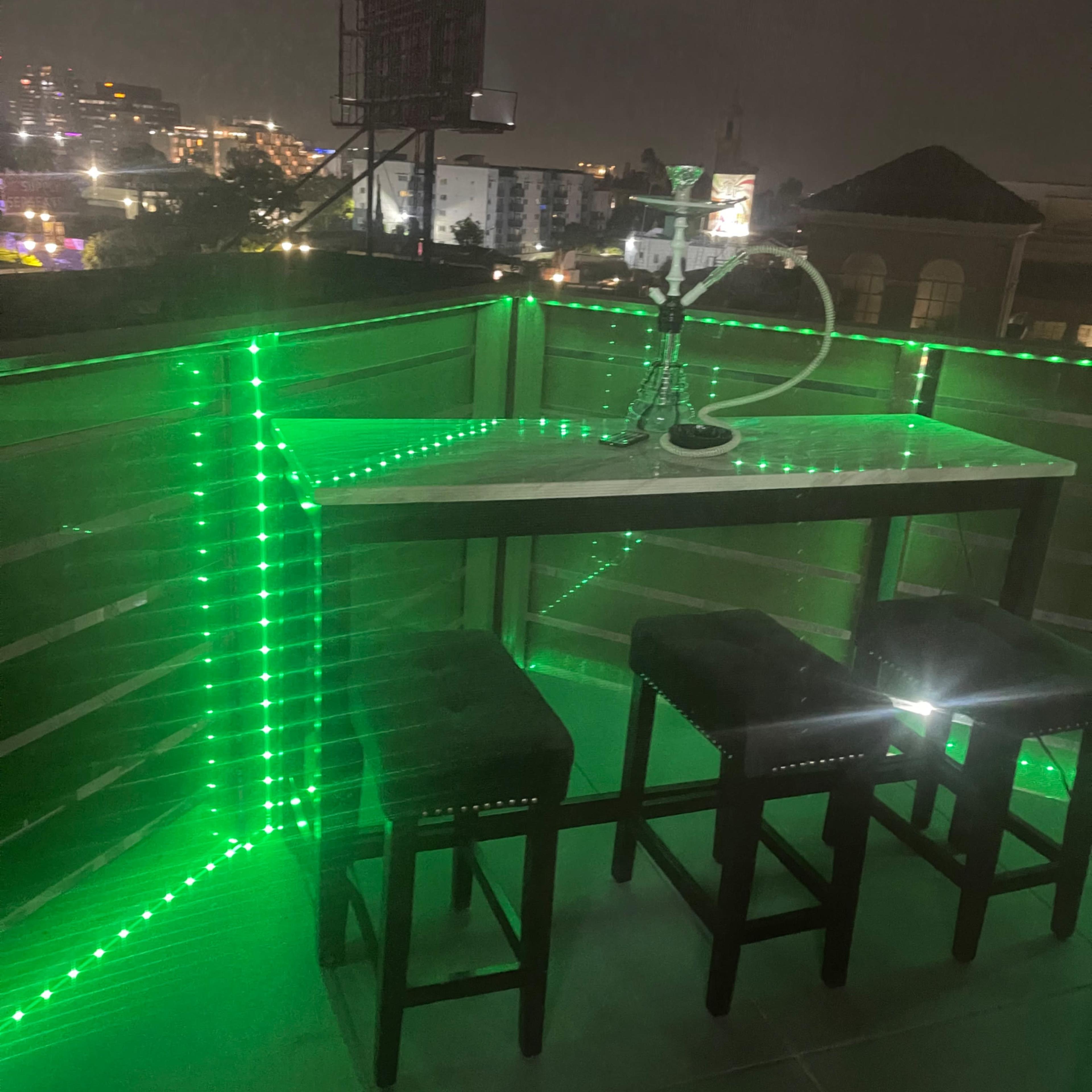 A modern balcony area features a table with three stools, illuminated by green LED lights, and a hookah on the table.