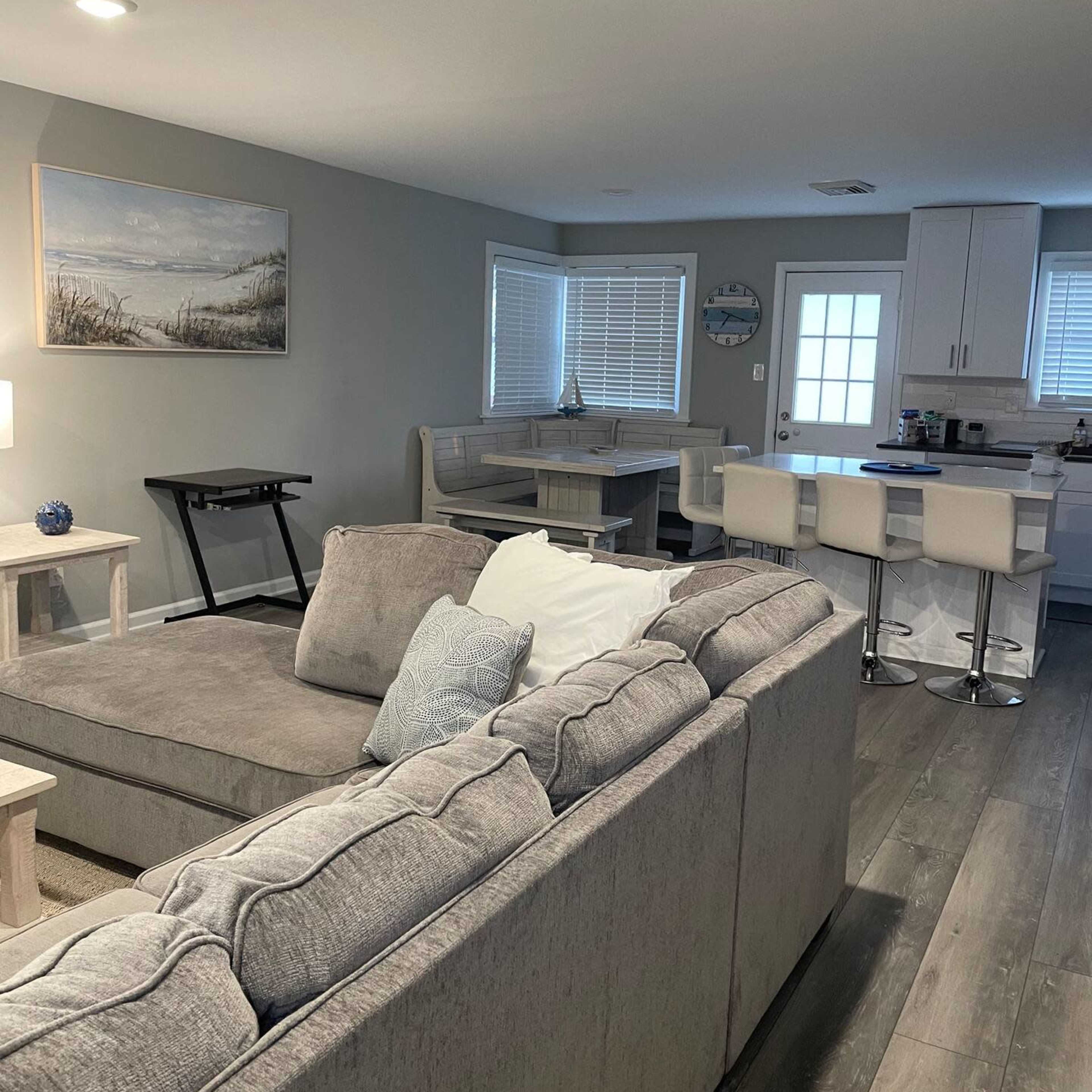 A gray sectional sofa faces a dining area with a table and chairs, while a kitchen is visible in the background with white cabinetry.