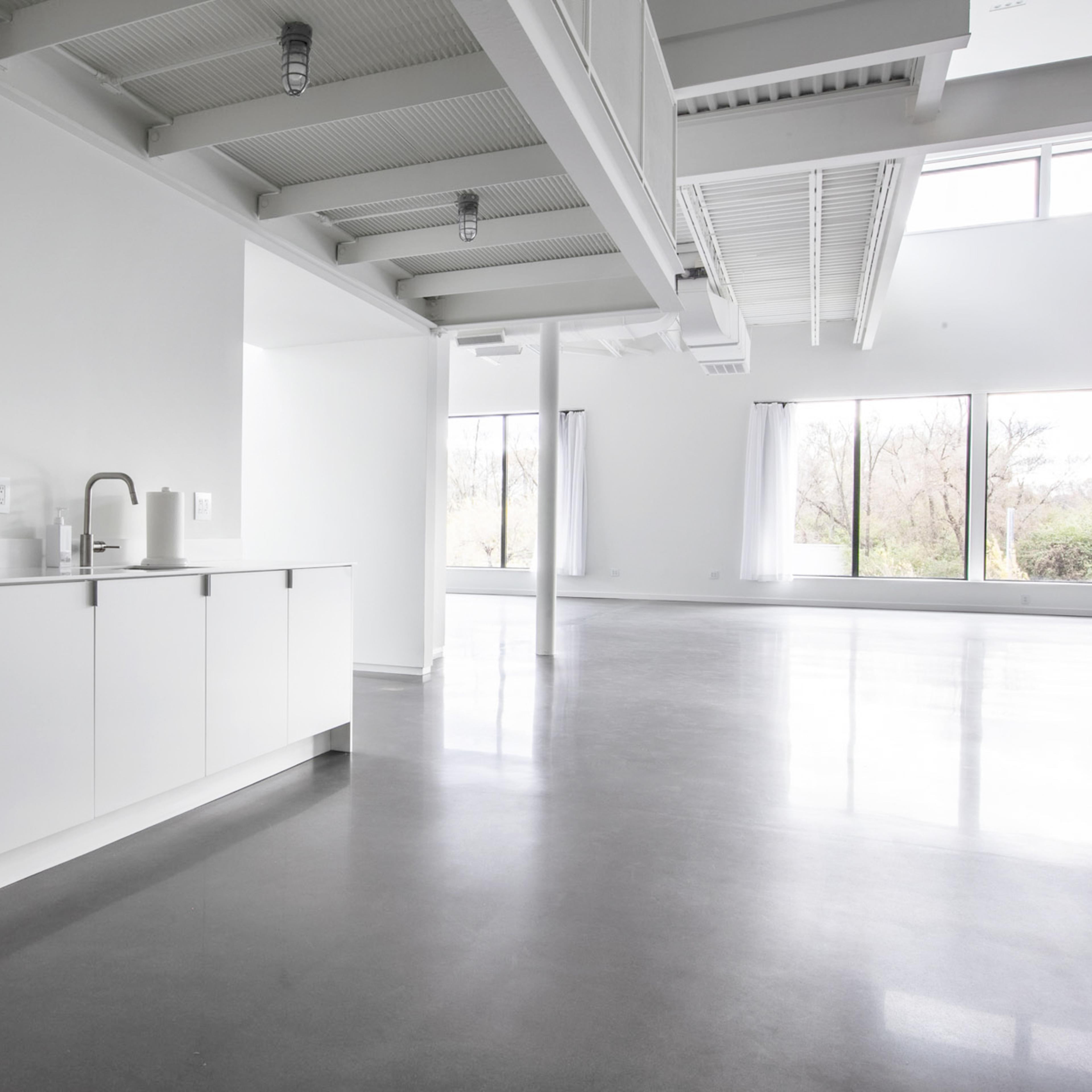 A minimalist, open-concept interior with a small kitchen area featuring a black refrigerator and a countertop, illuminated by large windows that provide natural light.