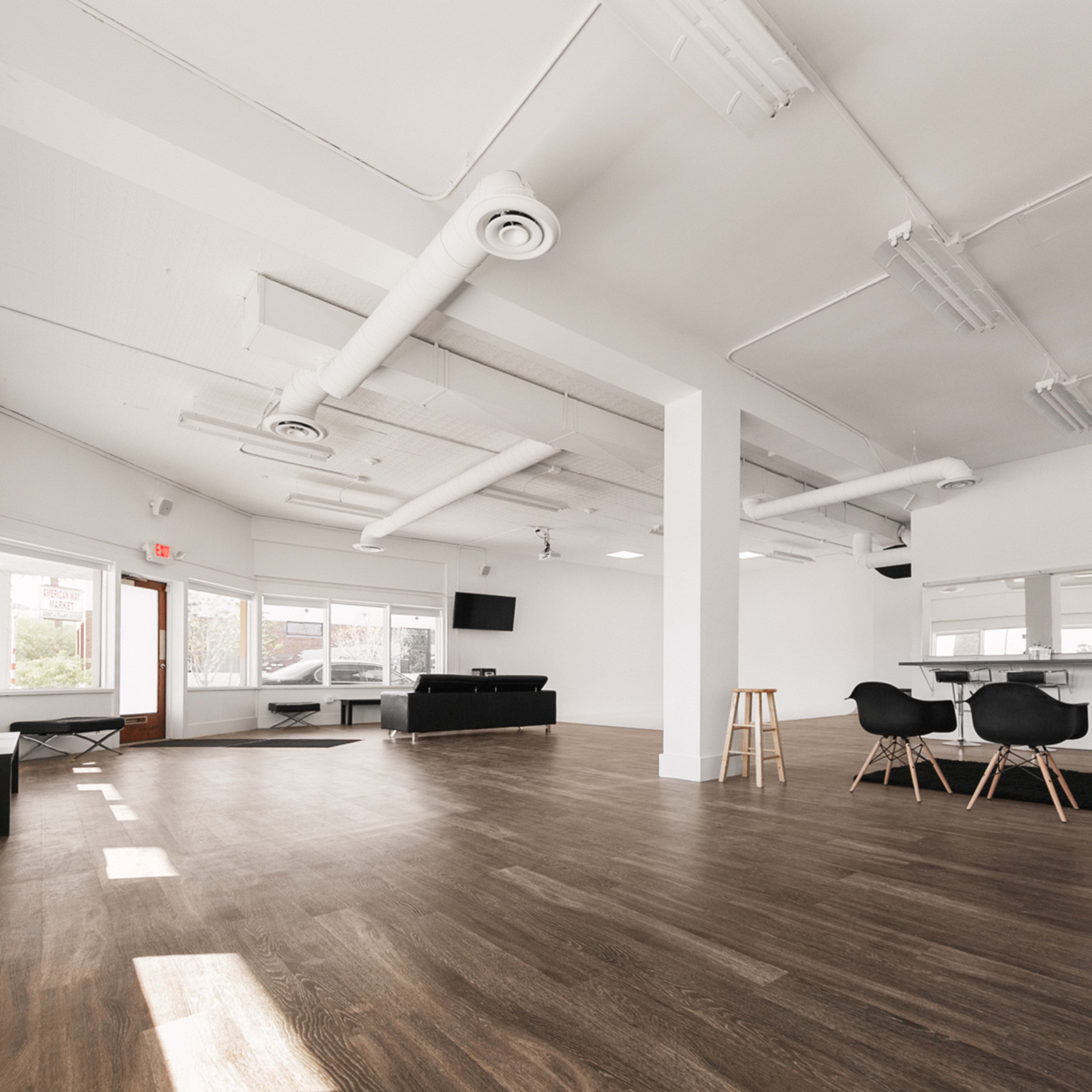 A spacious, minimalist room with large windows, wooden flooring, and a few pieces of furniture, including chairs and a stool.