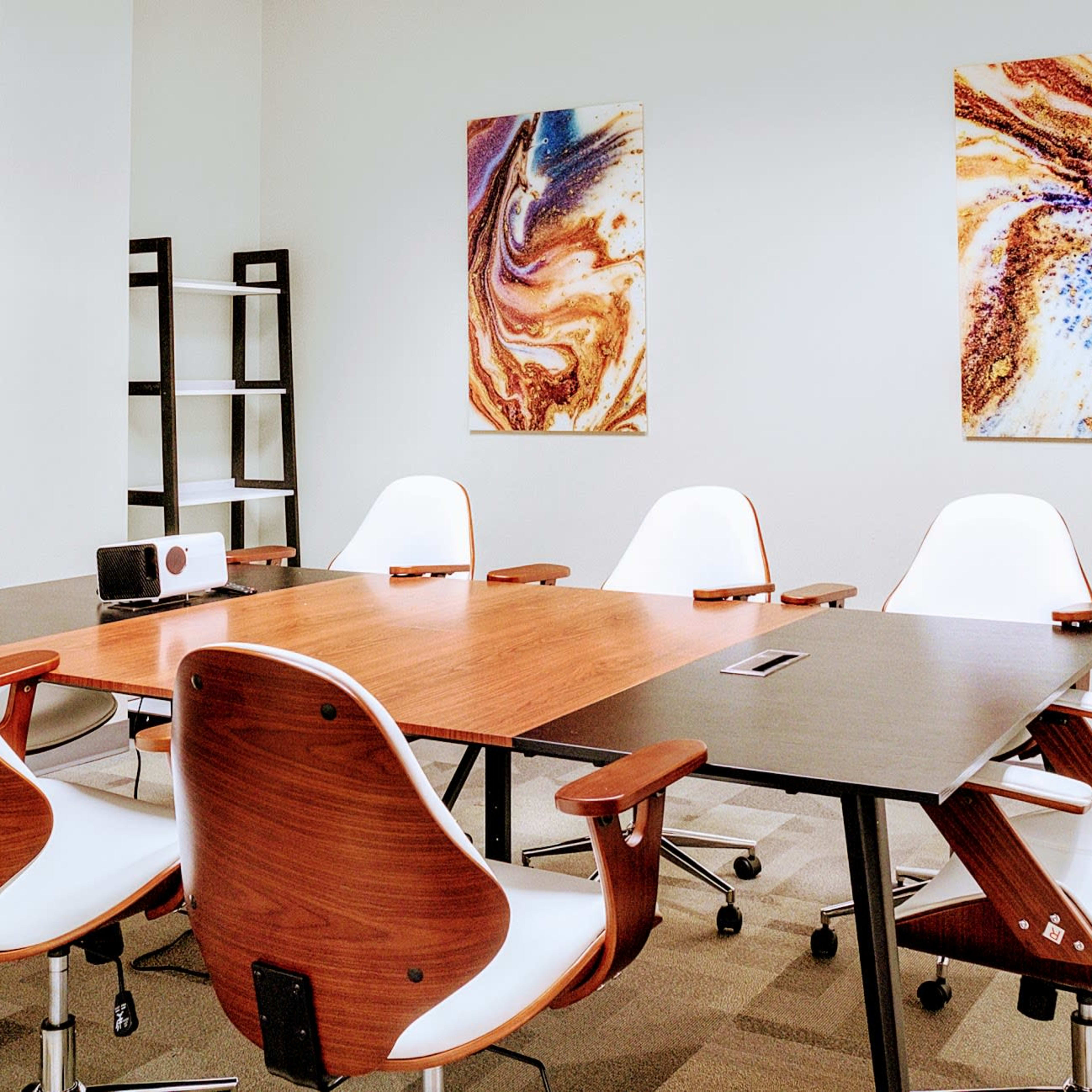 A conference room features a wooden table surrounded by white chairs, with abstract art on the walls.