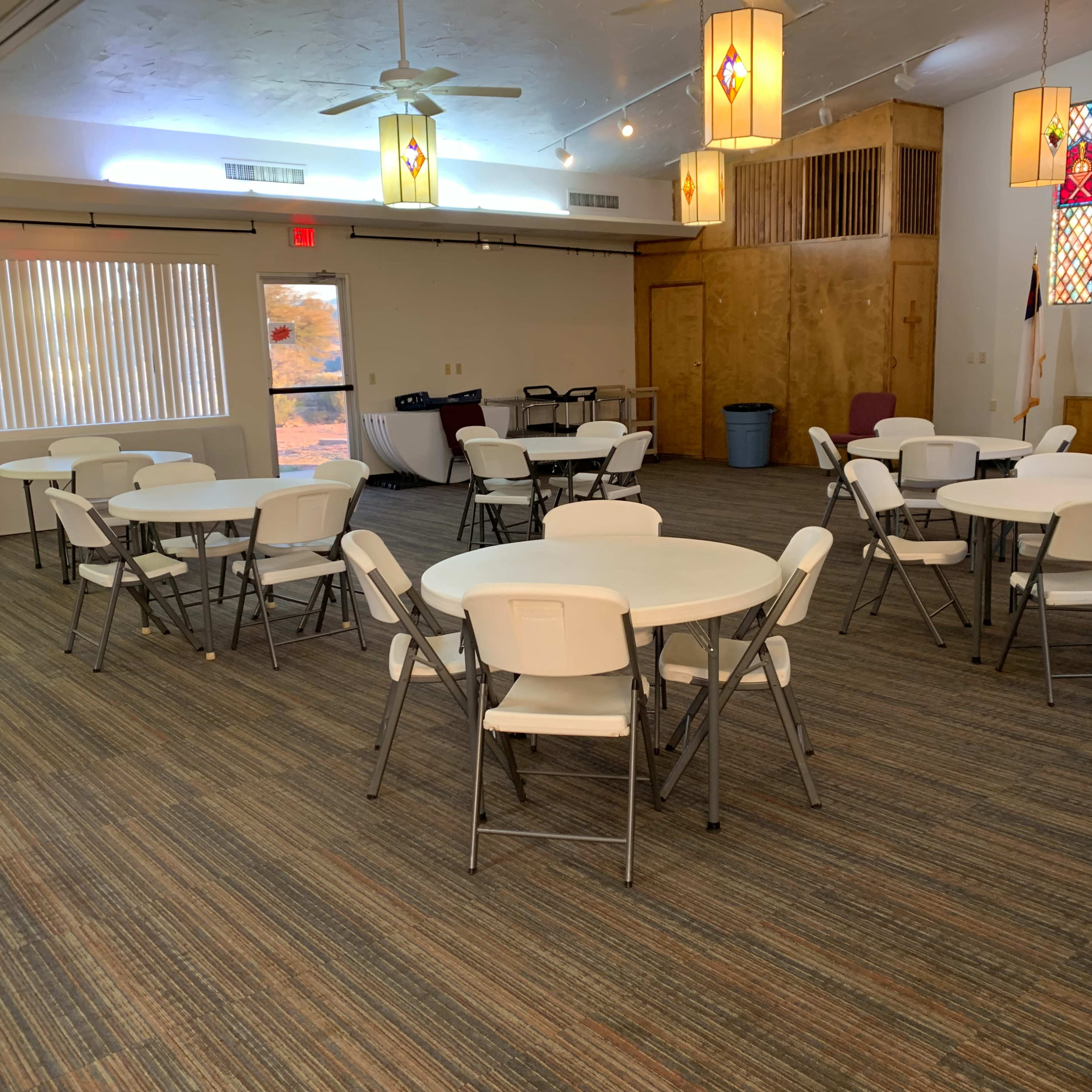 A spacious room with several round tables and white folding chairs, featuring stained glass windows and a wooden stage area.