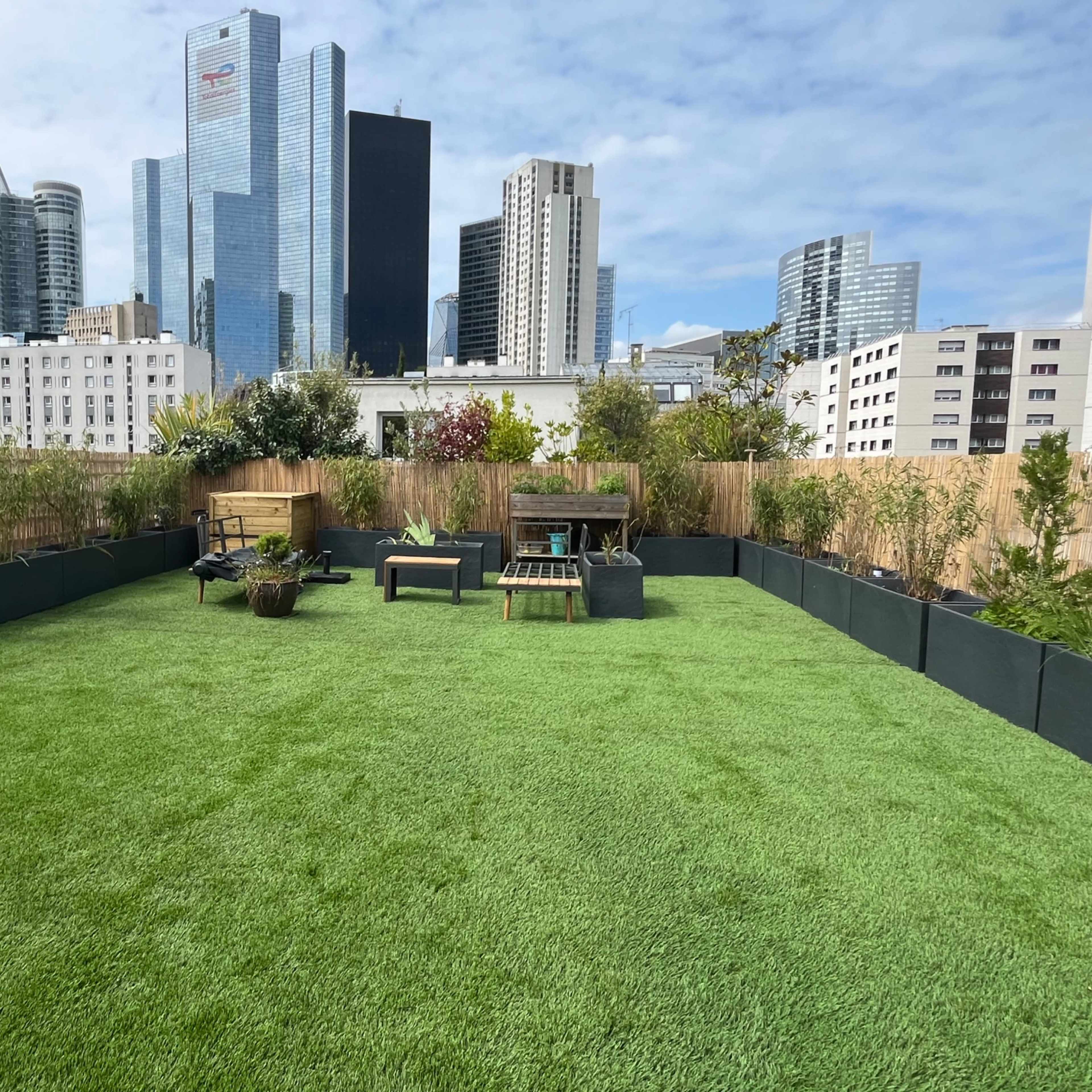A rooftop garden with artificial grass, featuring seating areas and surrounding greenery, set against a city skyline.