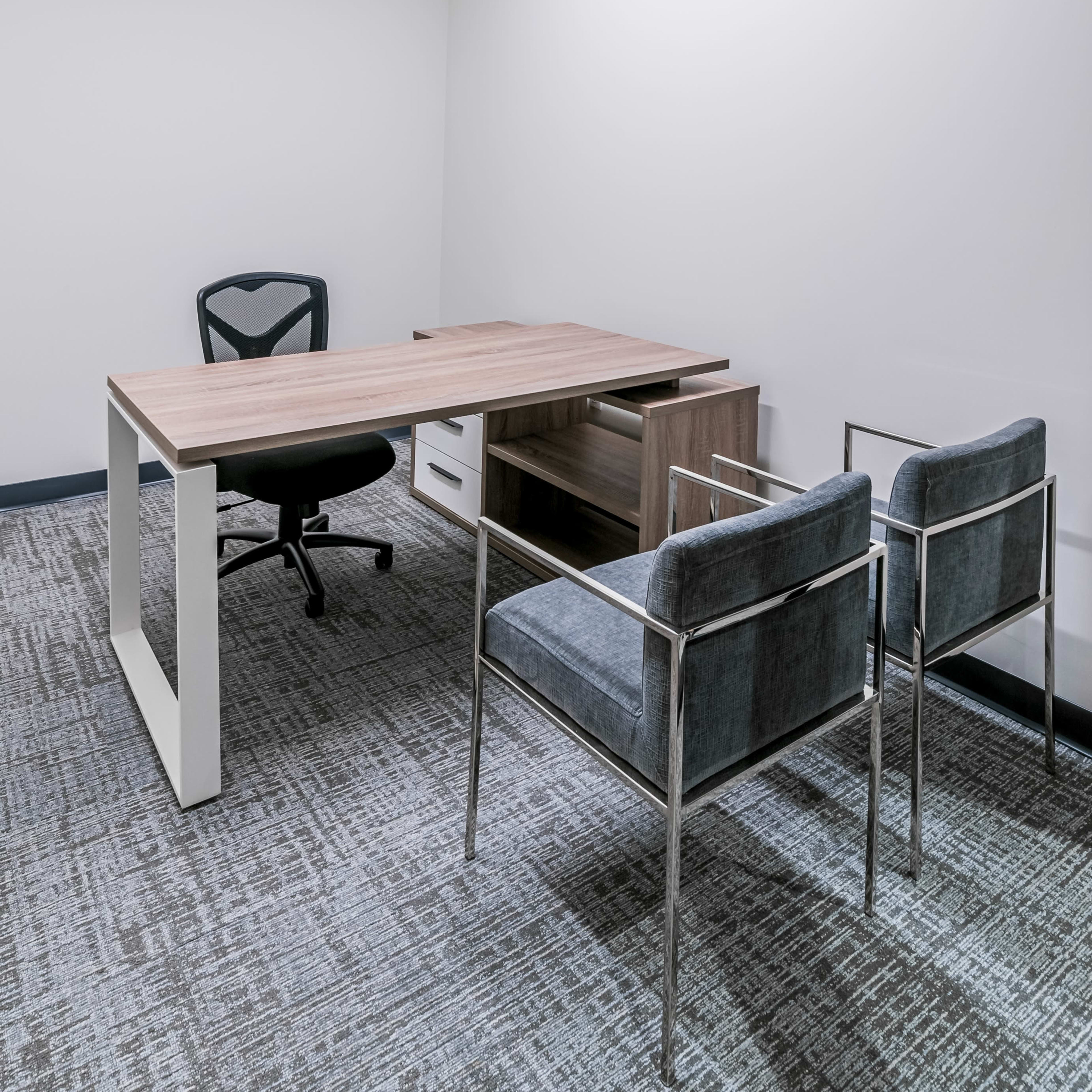 A modern office space features a wooden desk, a black office chair, and two gray upholstered chairs against a plain wall.