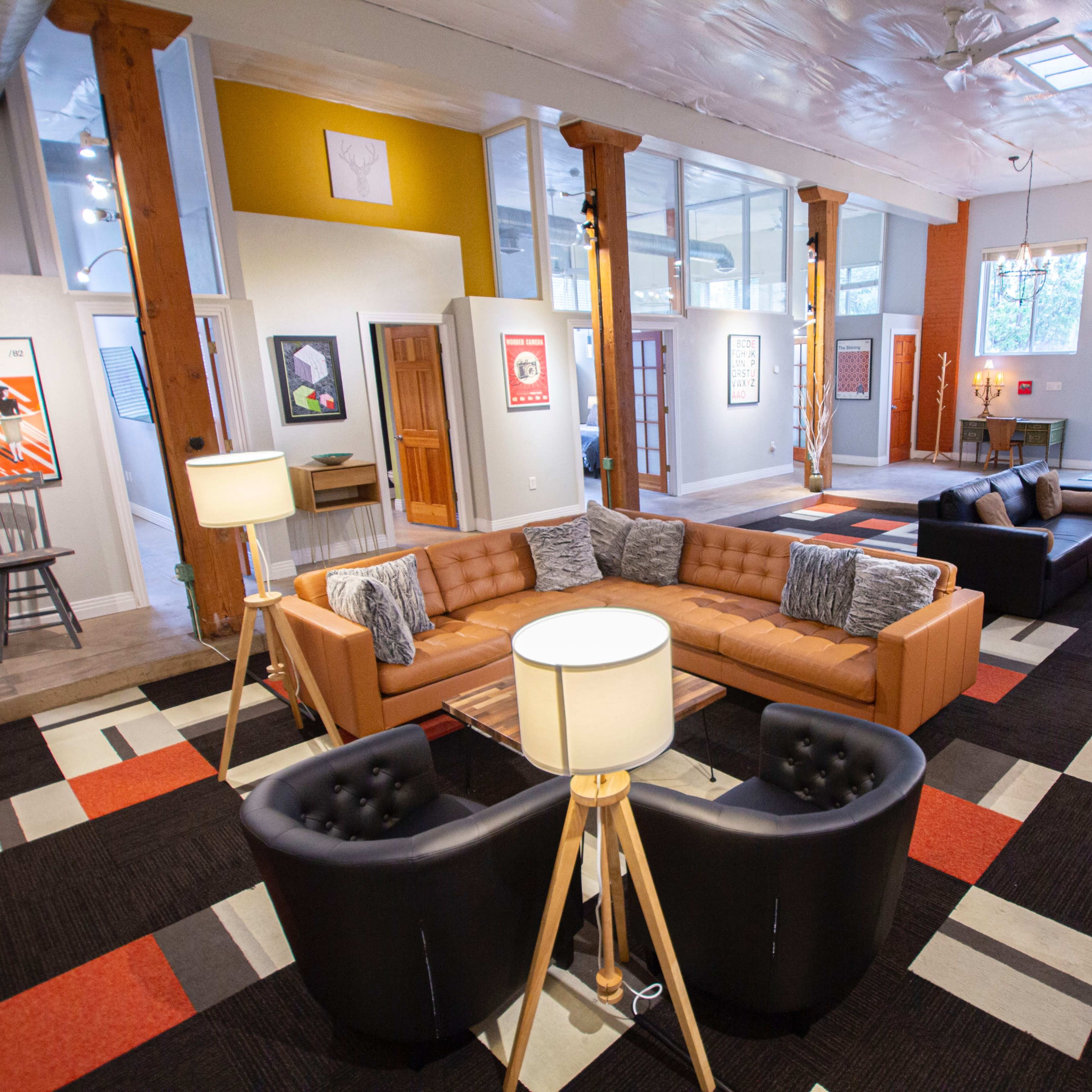 A spacious modern living room with a combination of a brown sectional sofa, black chairs, and colorful carpet, illuminated by lamps and natural light.