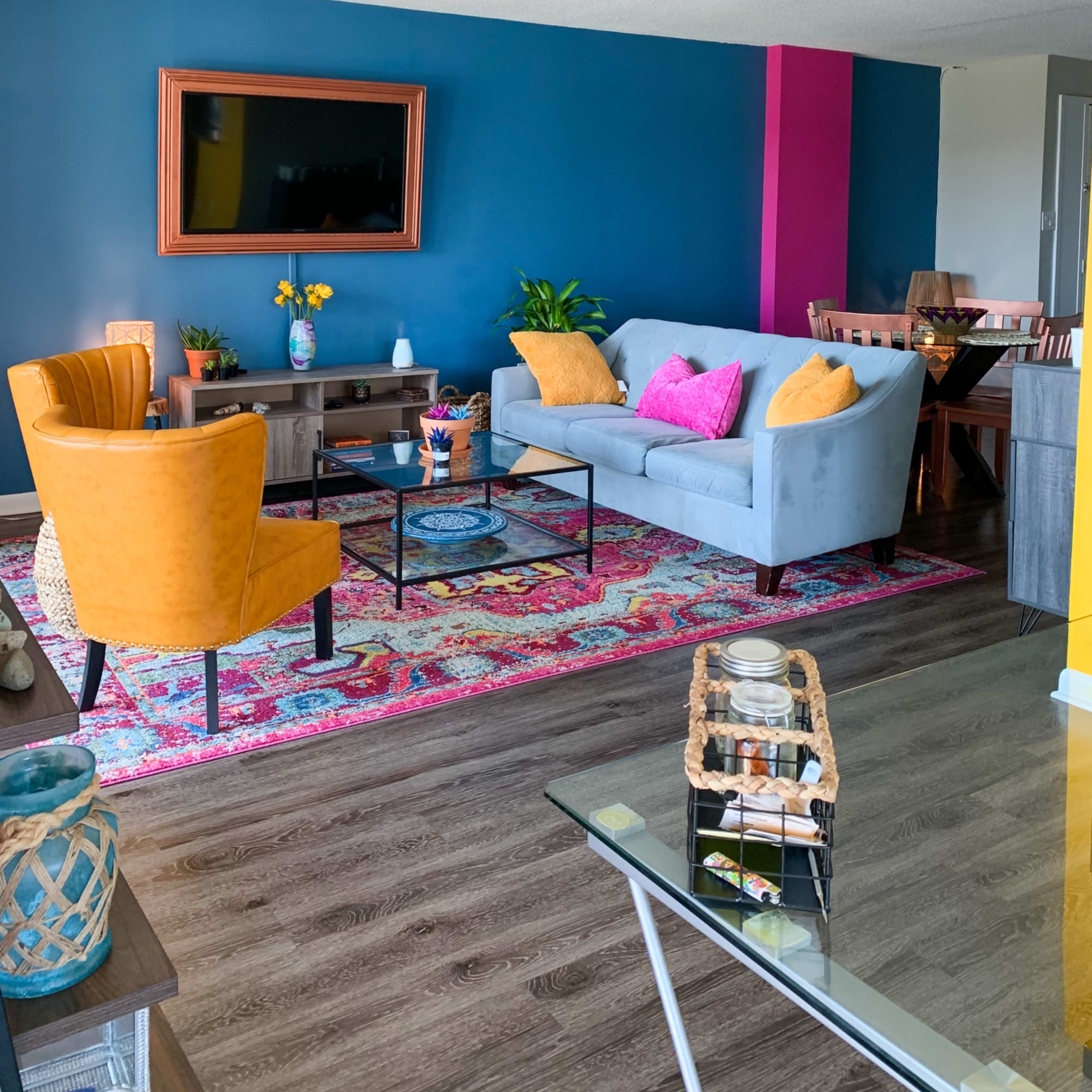 The living room features a blue accent wall, colorful decor, a gray sofa, two yellow armchairs, and a patterned area rug.