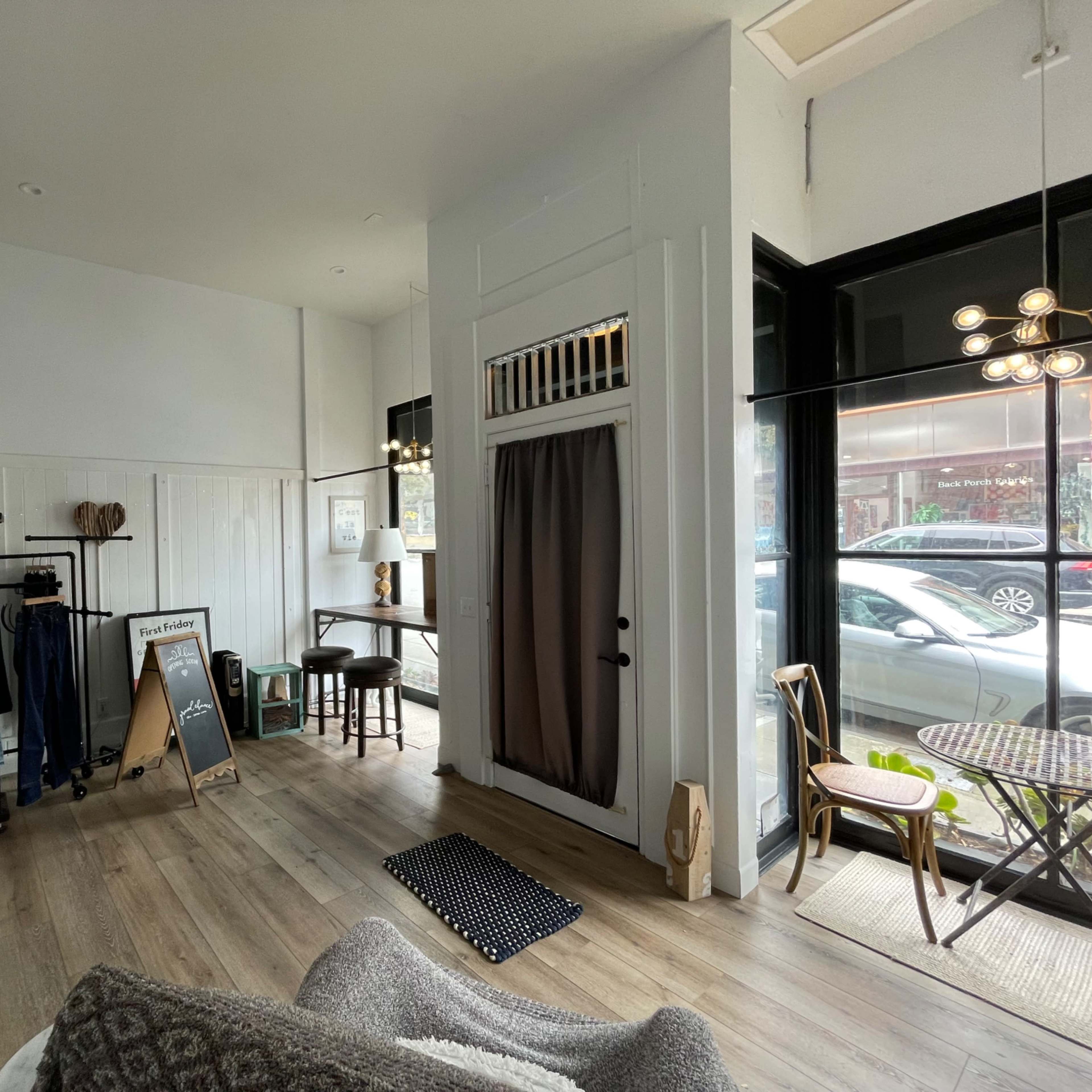 A brightly lit boutique interior with clothing racks, a small wooden table, and large windows facing the street.