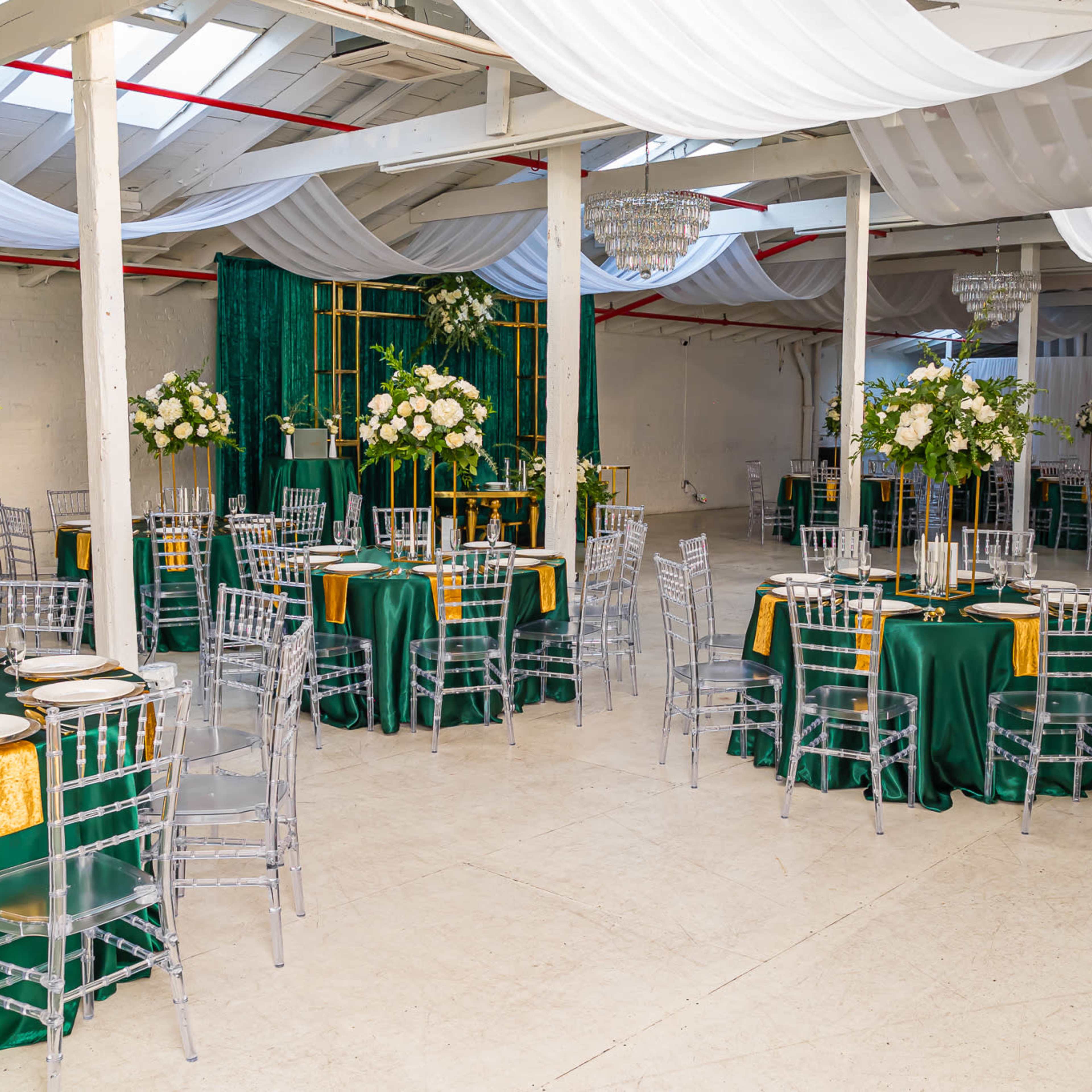 A spacious event venue set up with round tables covered in green tablecloths, adorned with floral centerpieces and elegant dining ware, under a canopy of draped fabric.