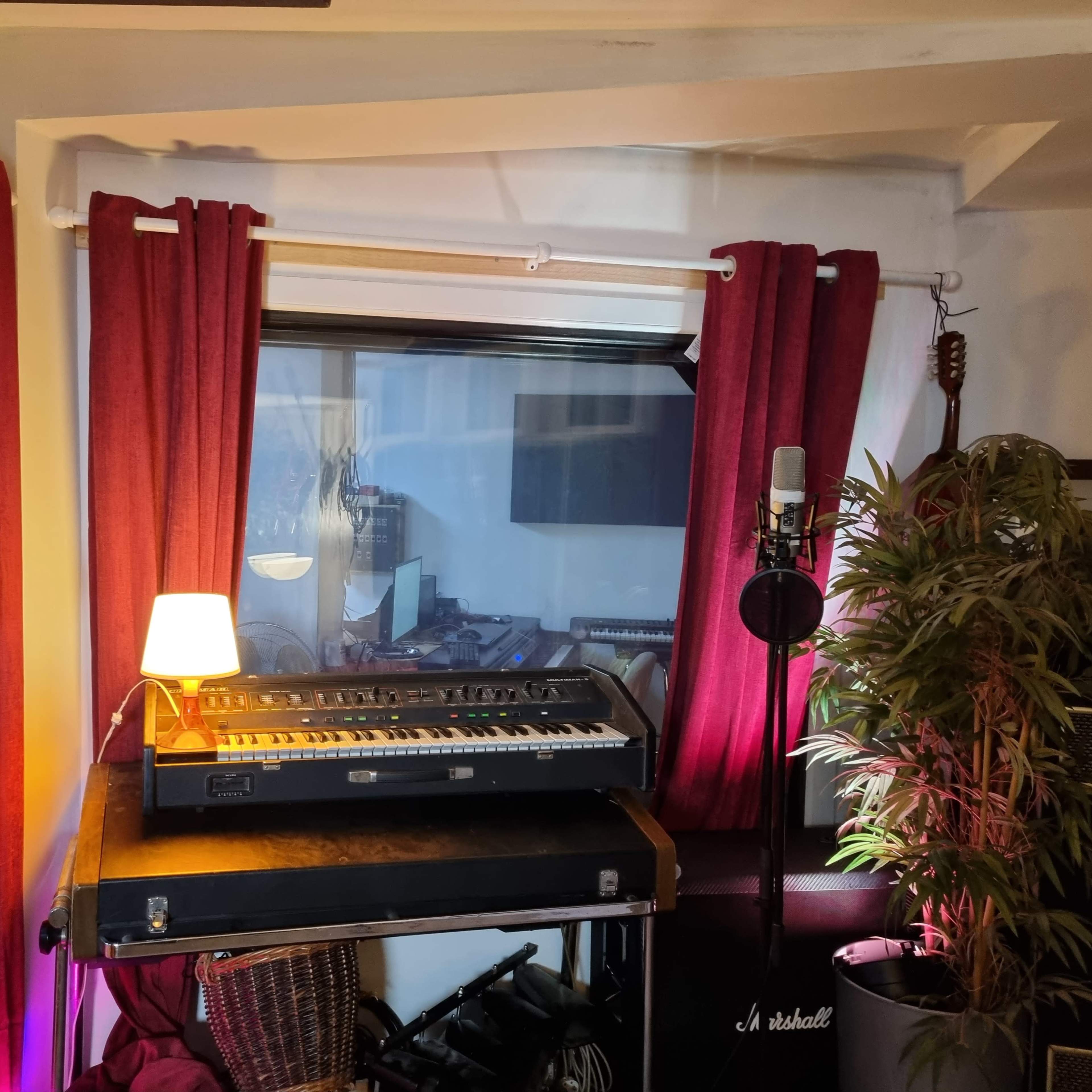 A small music studio corner with two microphones on stands, a lamp on a keyboard workstation, and red curtains framing a window.