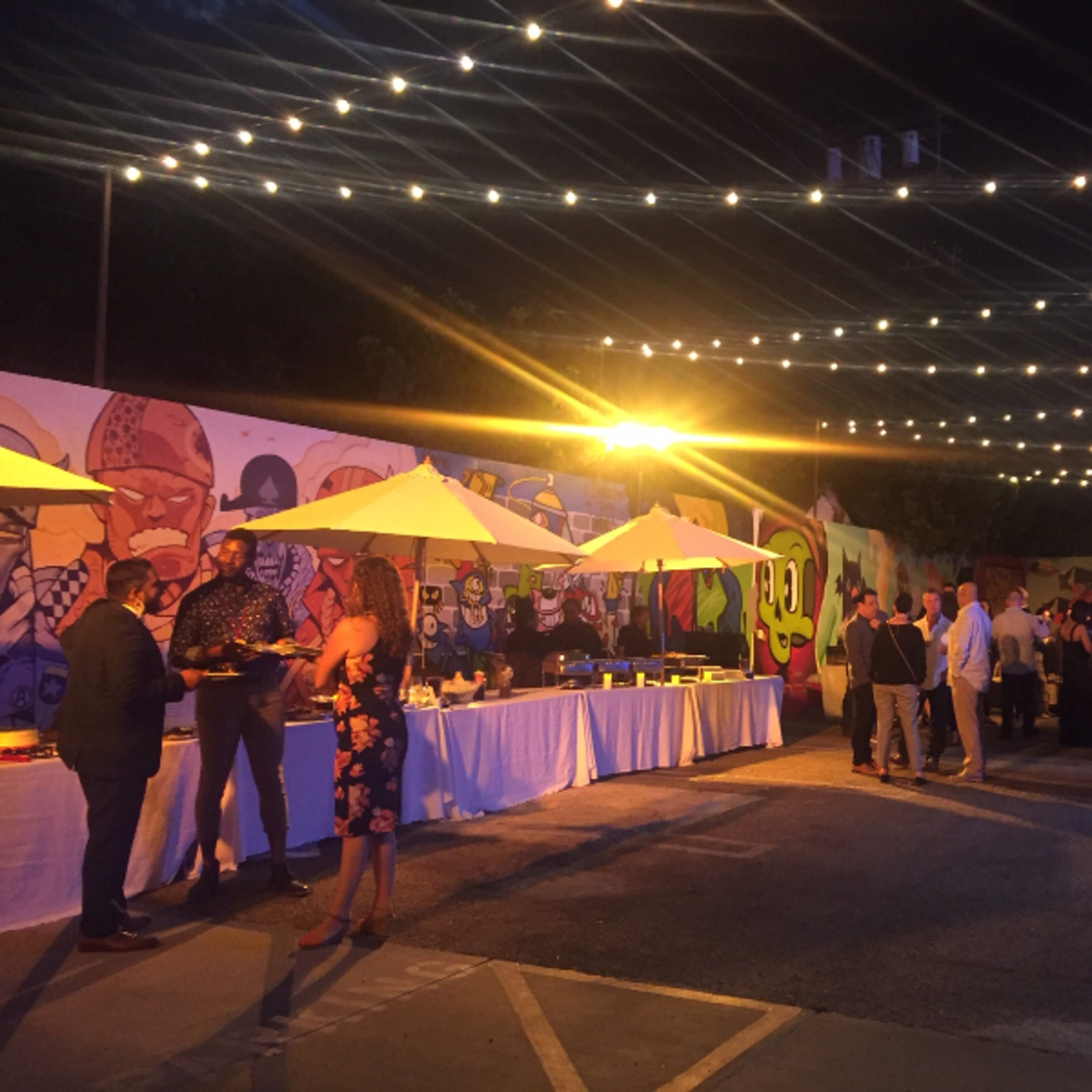 A lively outdoor event takes place under string lights, featuring tables with food and a colorful mural in the background.