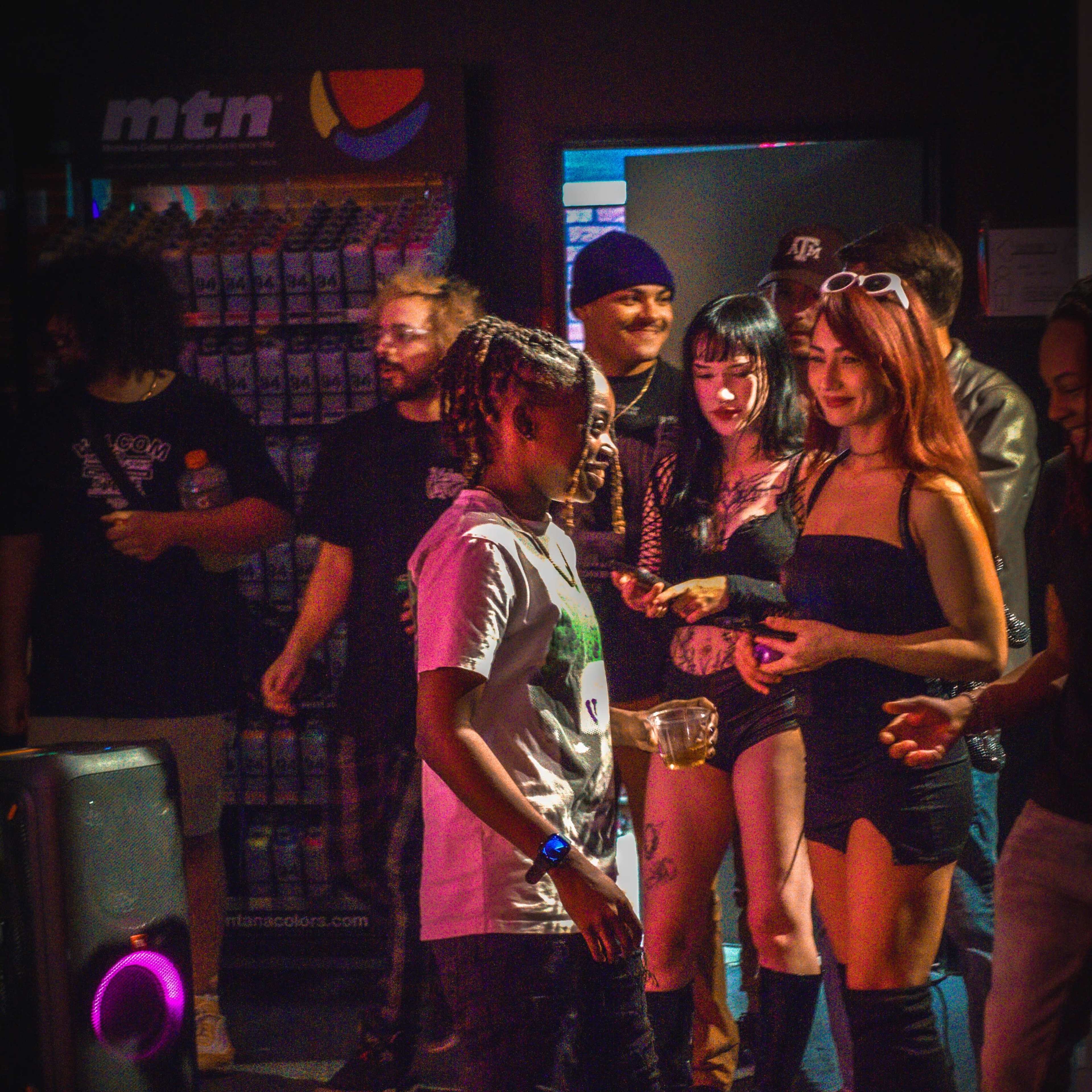 A group of people socializes in a dimly lit venue, with a mix of individuals dressed in various styles, including one person in a graphic t-shirt and another in a black outfit.