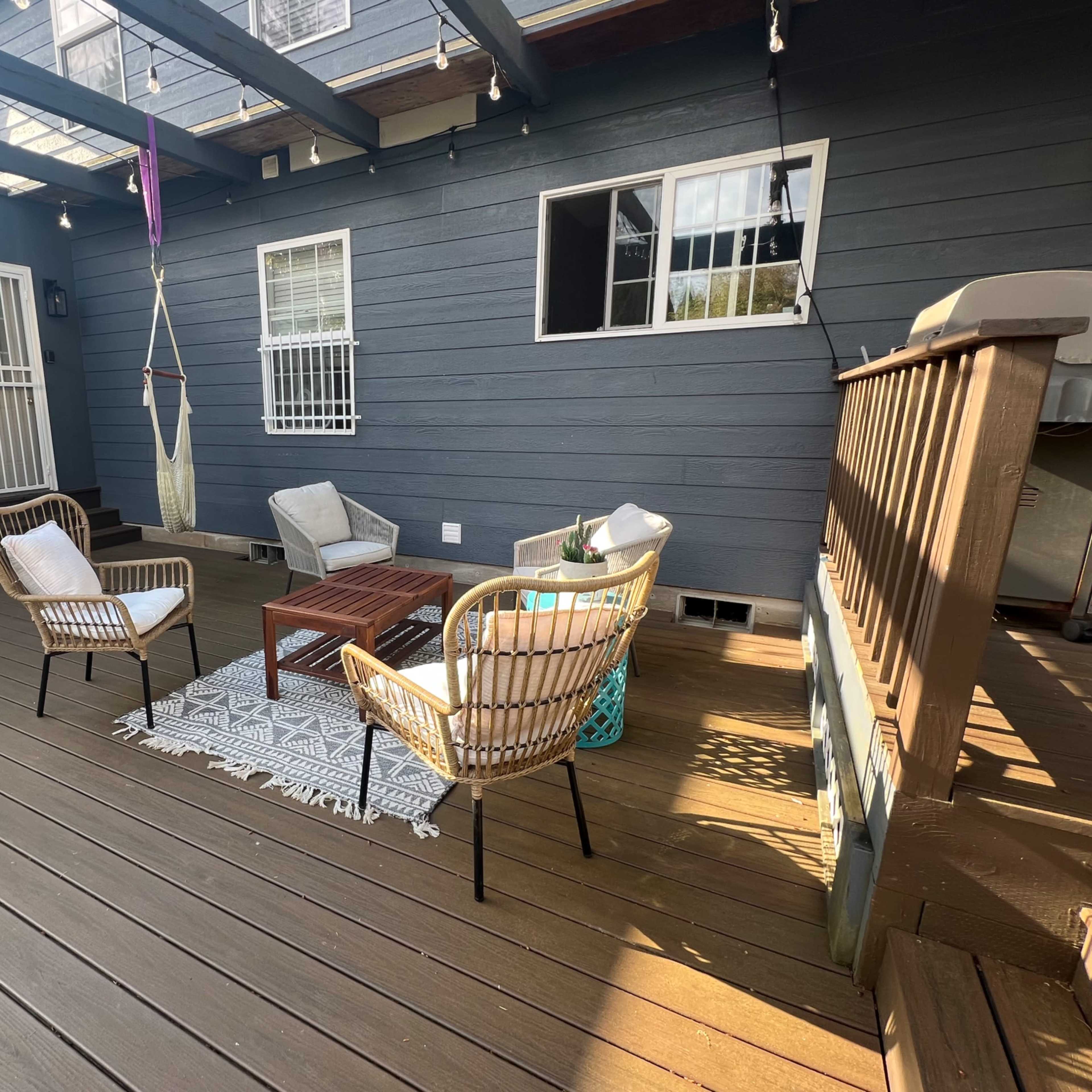 A deck area with a coffee table surrounded by four chairs and a hammock, set against a wall of blue wooden siding and large windows.