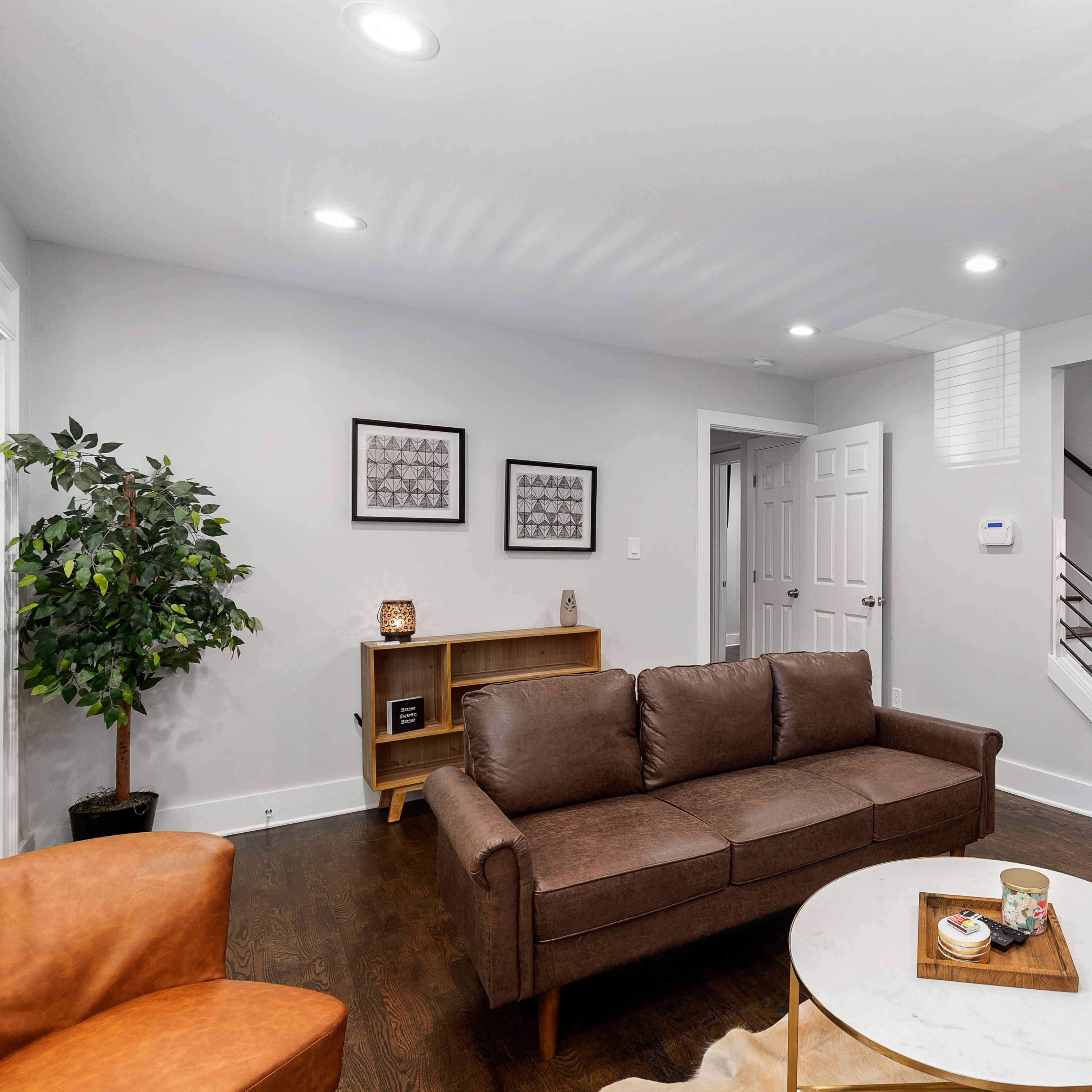 A modern living room with a brown sofa, a round coffee table, and a bookshelf, featuring a staircase and a dining area in the background.